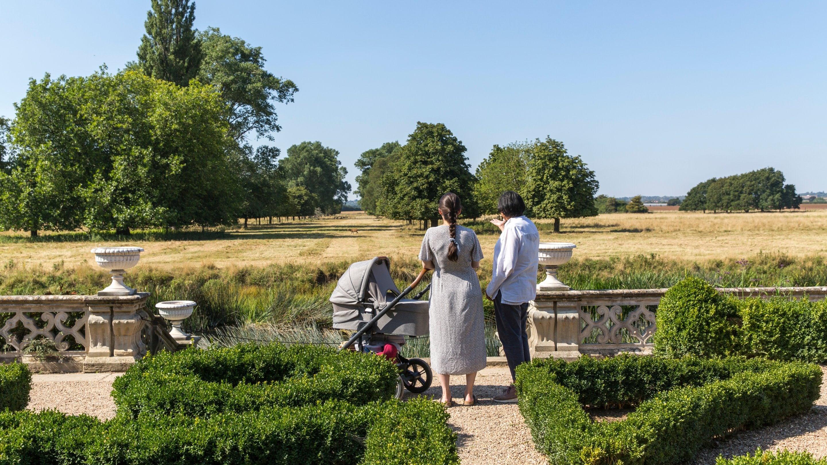 A young mother and her mother (grandmother) stand next to a pram between small box hedges that form a pattern on the parterre. They are looking out across the river in front of them and to the parkland in the far ground which is looking slightly parched in the summer weather. Blue skies and sunshine are above.