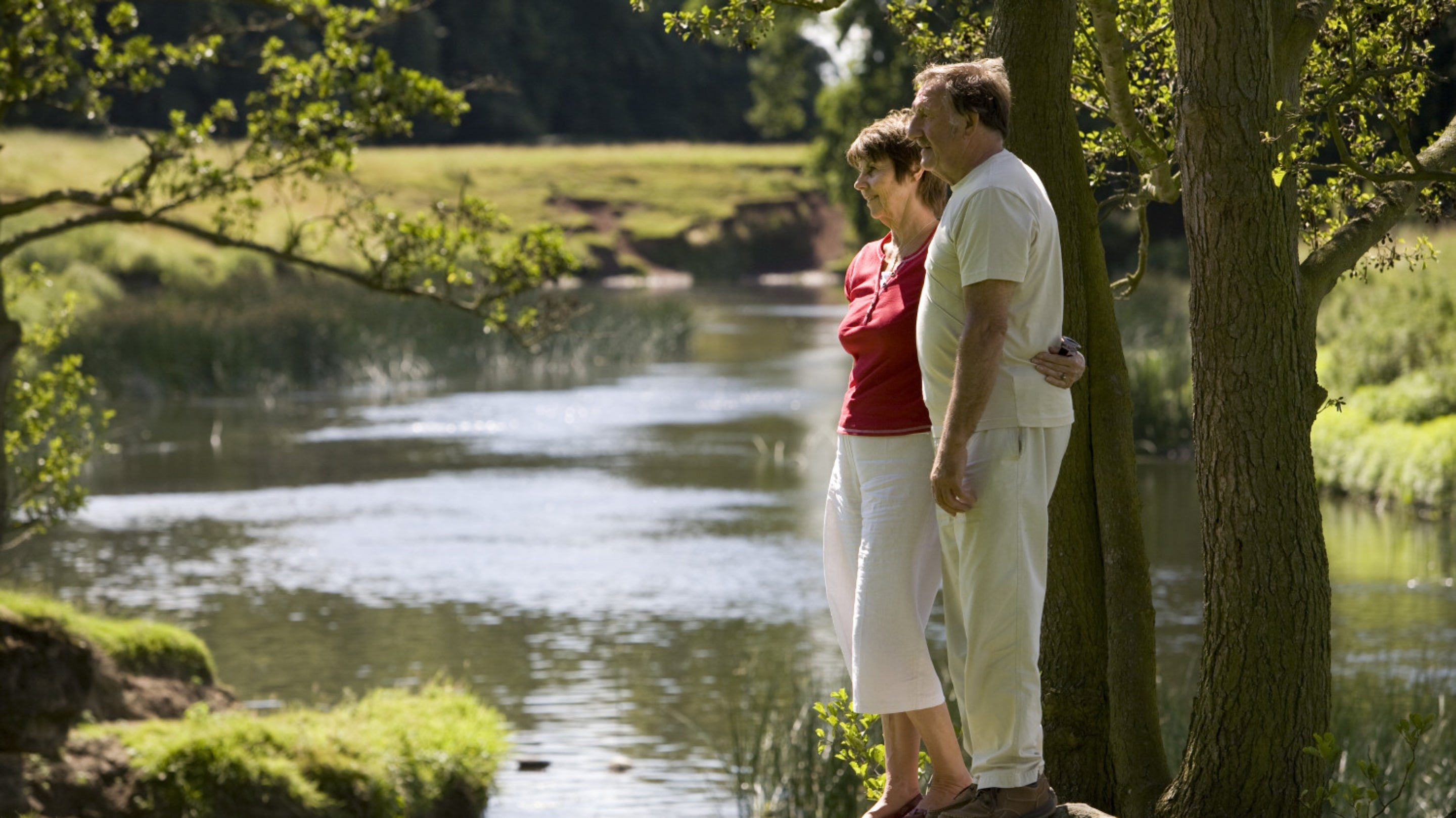 A couple holding hands and posing for a photo in front of the River Avon at Charlecote Park on a sunny day