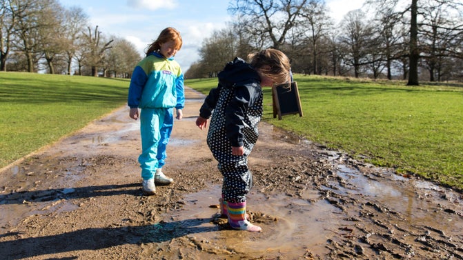 Two young children in full waterproof jump suits, splashing in muddy puddles against a background of blue skies.