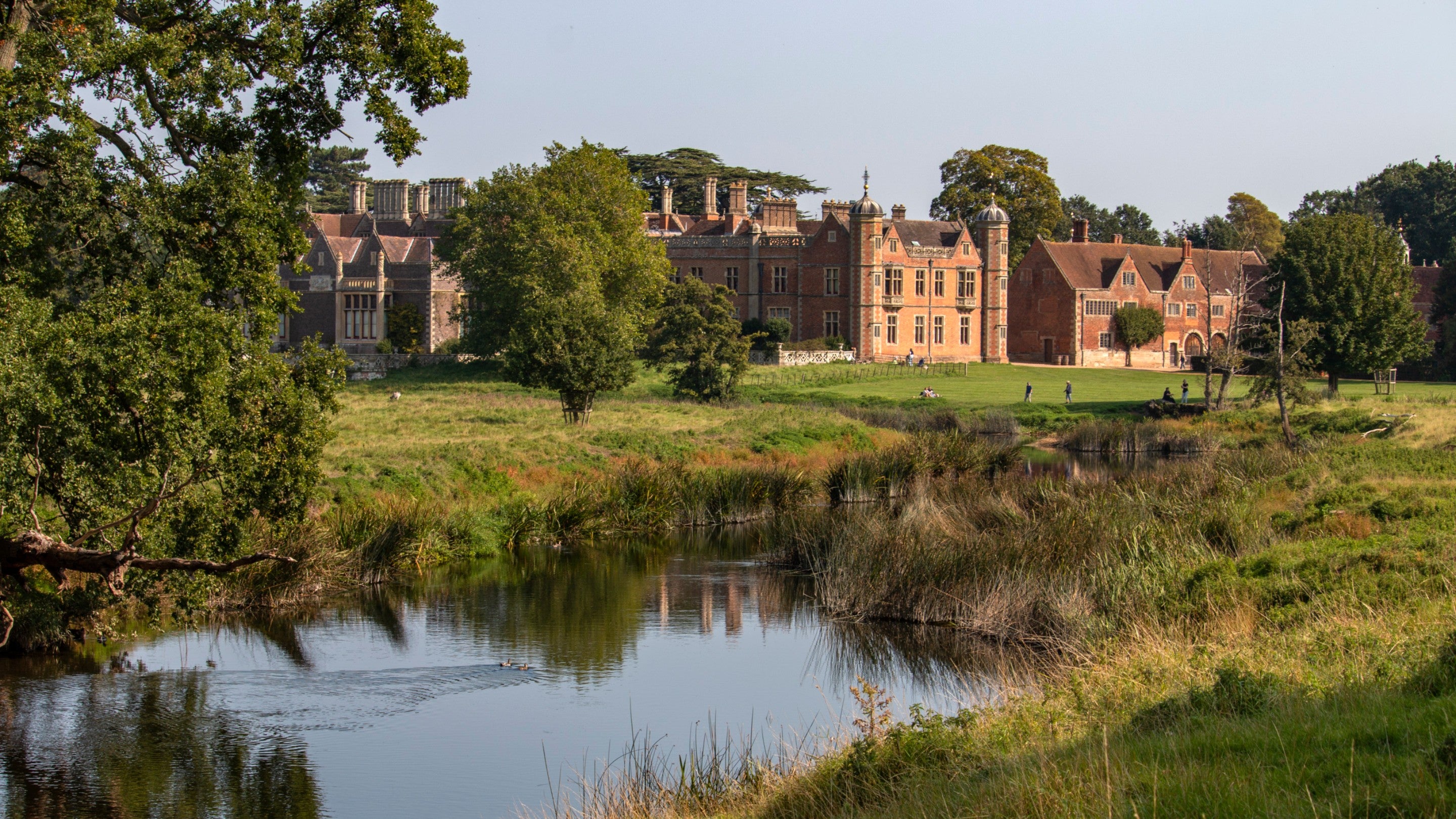 A view of the house at Charlecote Park from across the River Avon on a sunny day, with greenery on the banks and trees in front of the house