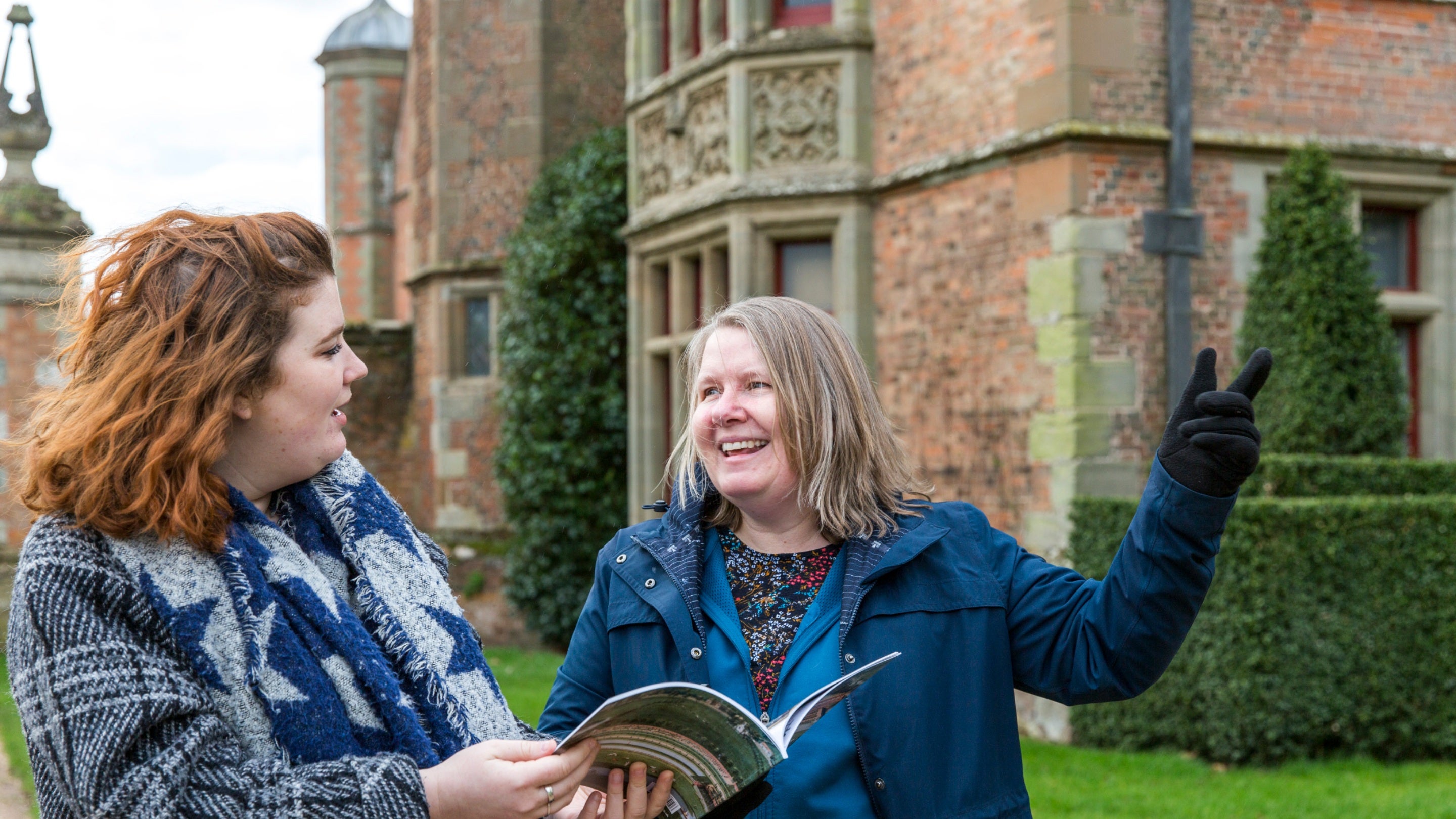 Two visitors in winter wear outside looking at a guidebook with parts of the red brick Charlecote Park behind