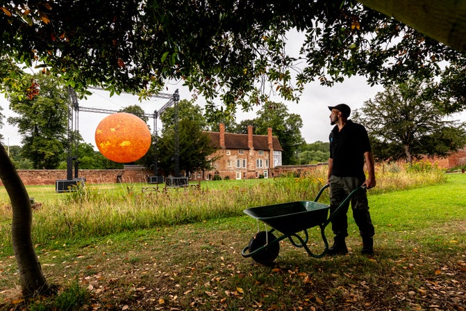 A gardener with his wheelbarrow looks over towards the bright yellow-red of Helios, the art installation of the sun, with the Charterhouse nestled on its right hand side