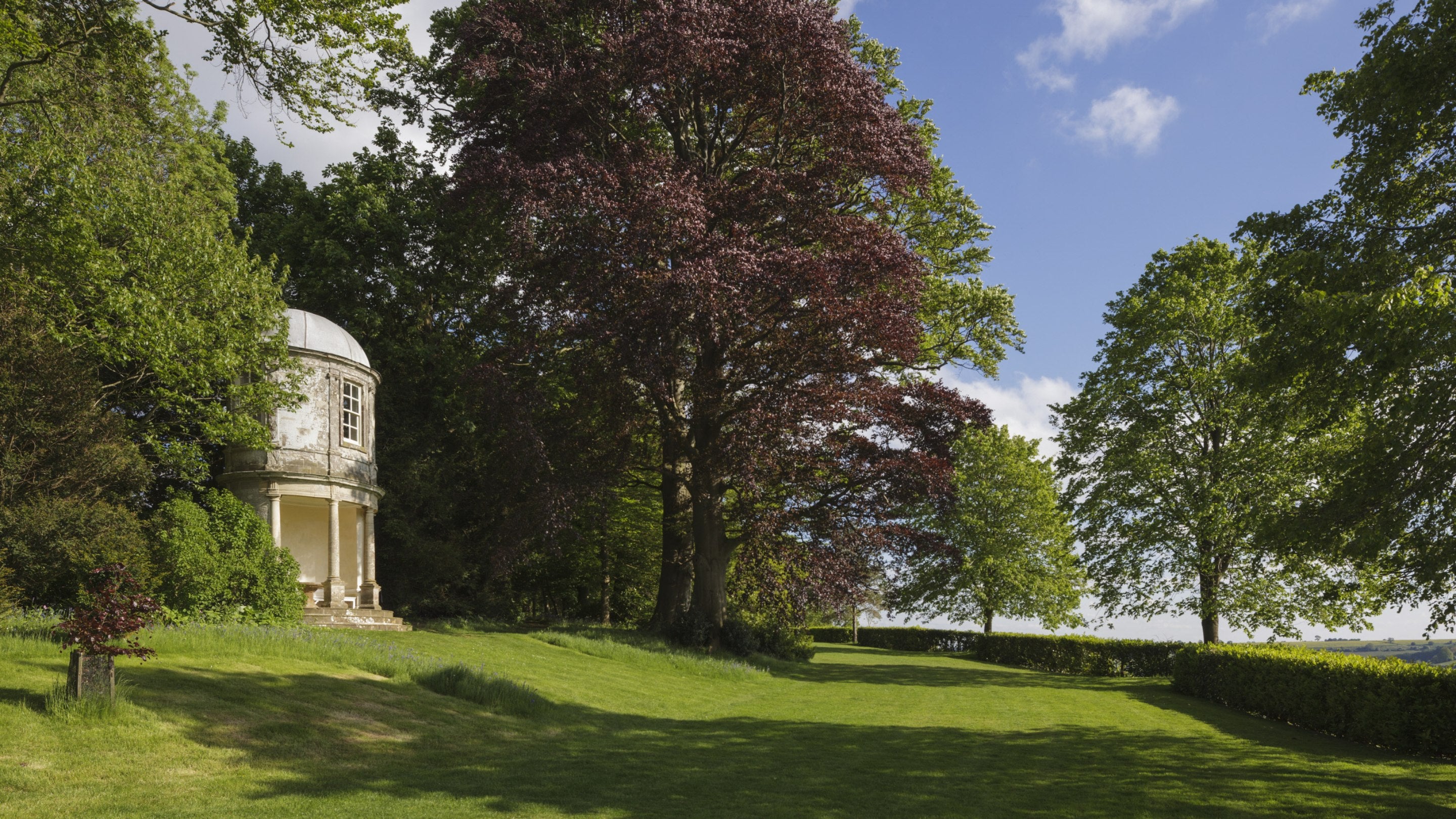 A round temple in the landscape garden at Farnborough Hall, surrounded by trees and lawns on a sunny day