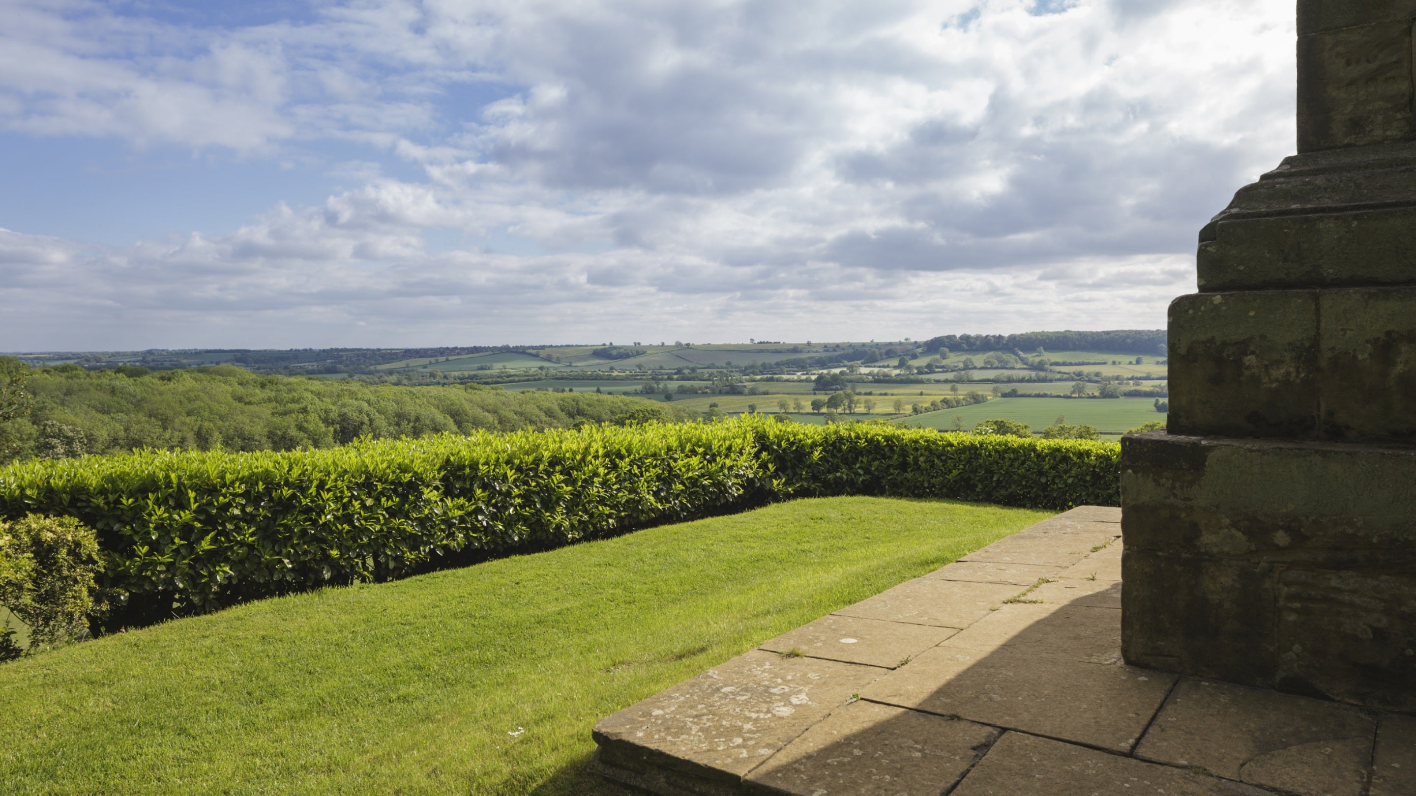 A view of the landscape from beside the obelisk at Farnborough Hall, with long-reaching views of the countryside beyond a neat hedge, a glimpse of the obelisk visible on the right