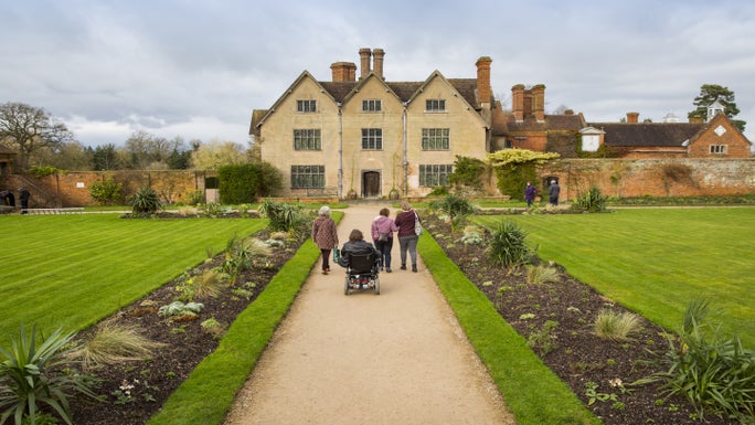 Four visitors approaching Packwood House along garden path with flowerbeds and lawns either side