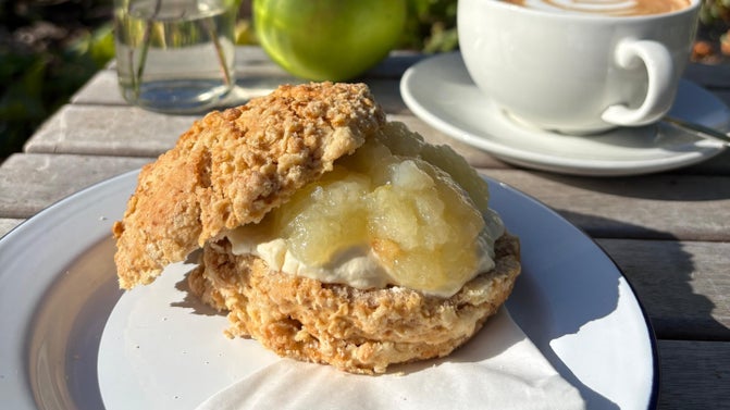 An apple crumble scone with apple compote and whipped custard on a plate, with a cup of coffee behind, at Packwood House, Warwickshire