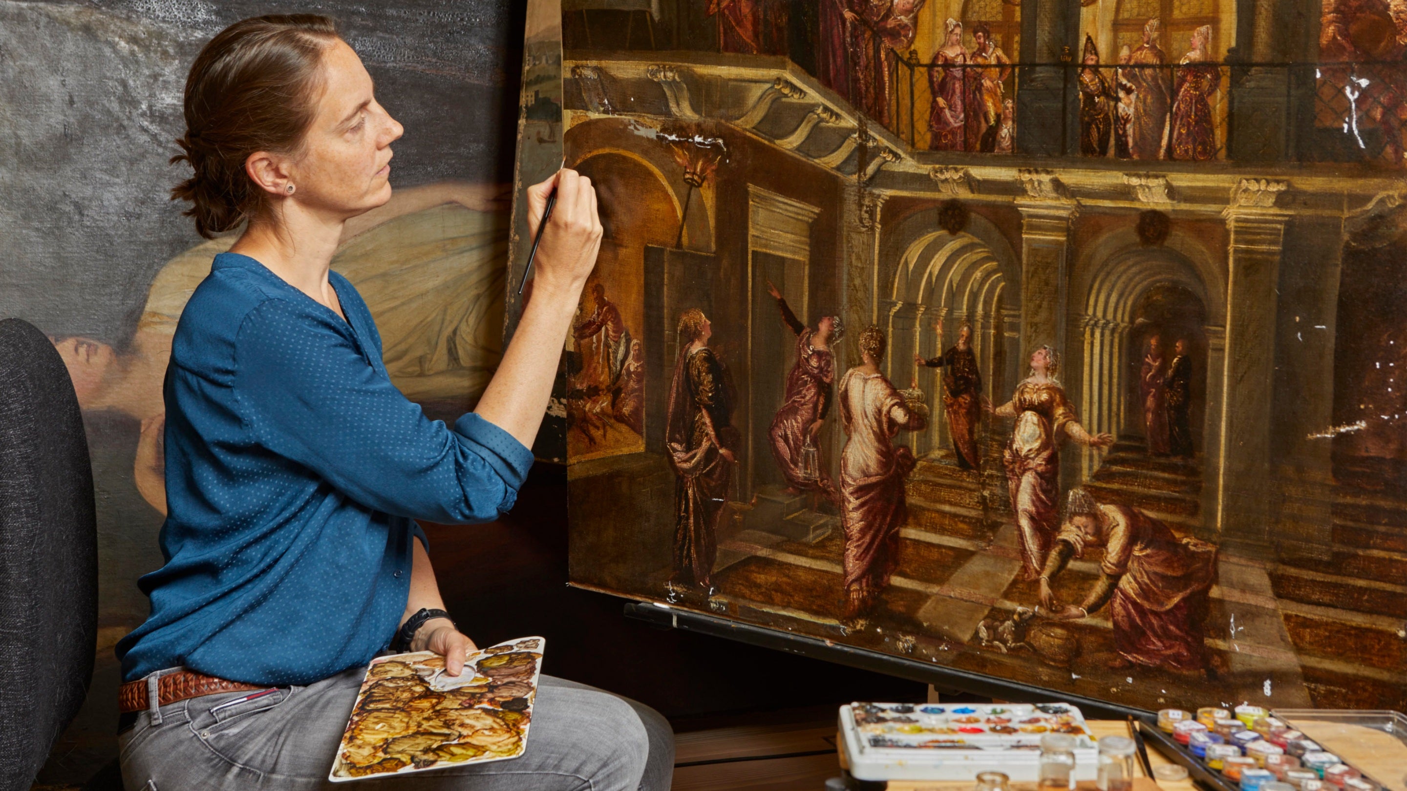 Female conservator sits in front of the painting doing some touch ups near the arches section. She holds a palette and uses a fine brush and has a mixture of coloured paints beside her.