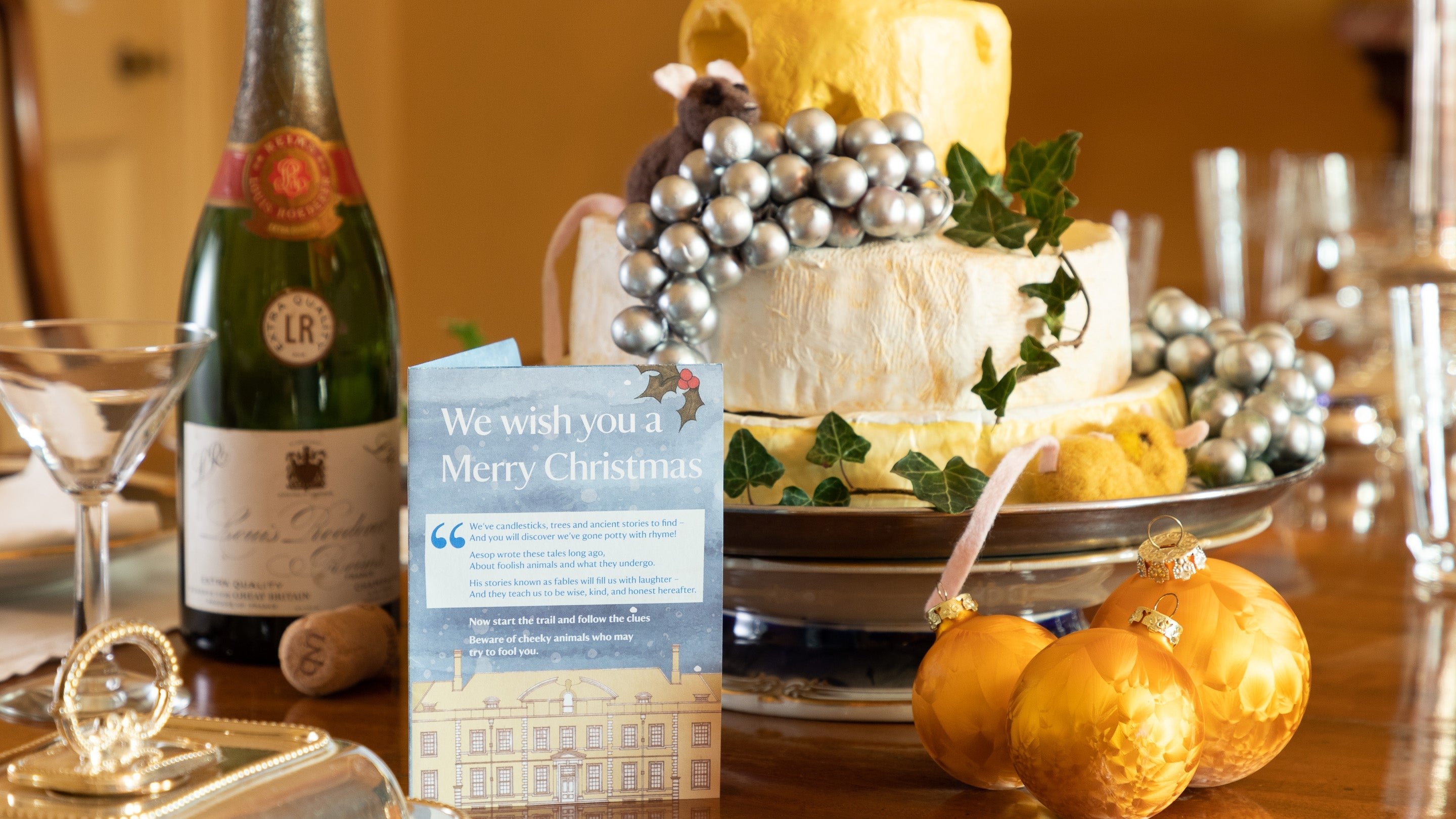 A Christmas dining table at Upton House and Garden, Warwickshire, is laid with a central platter of cheeses, decorated with silver grapes and a little mouse. Also on the table are a bottle of Champagne, glasses, a Christmas card from the National Trust saying 'We wish you a merry Christmas', and golden baubles.