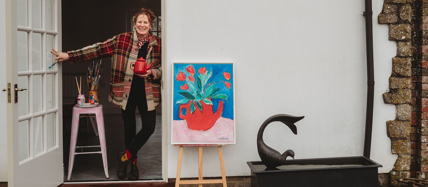 Artist Georgie Weedon posing in the doorway of the Racquets Court in Avebury Manor Garden, next to a colourful painting of a red jug containing bright red flowers against a blue backgrounfd.