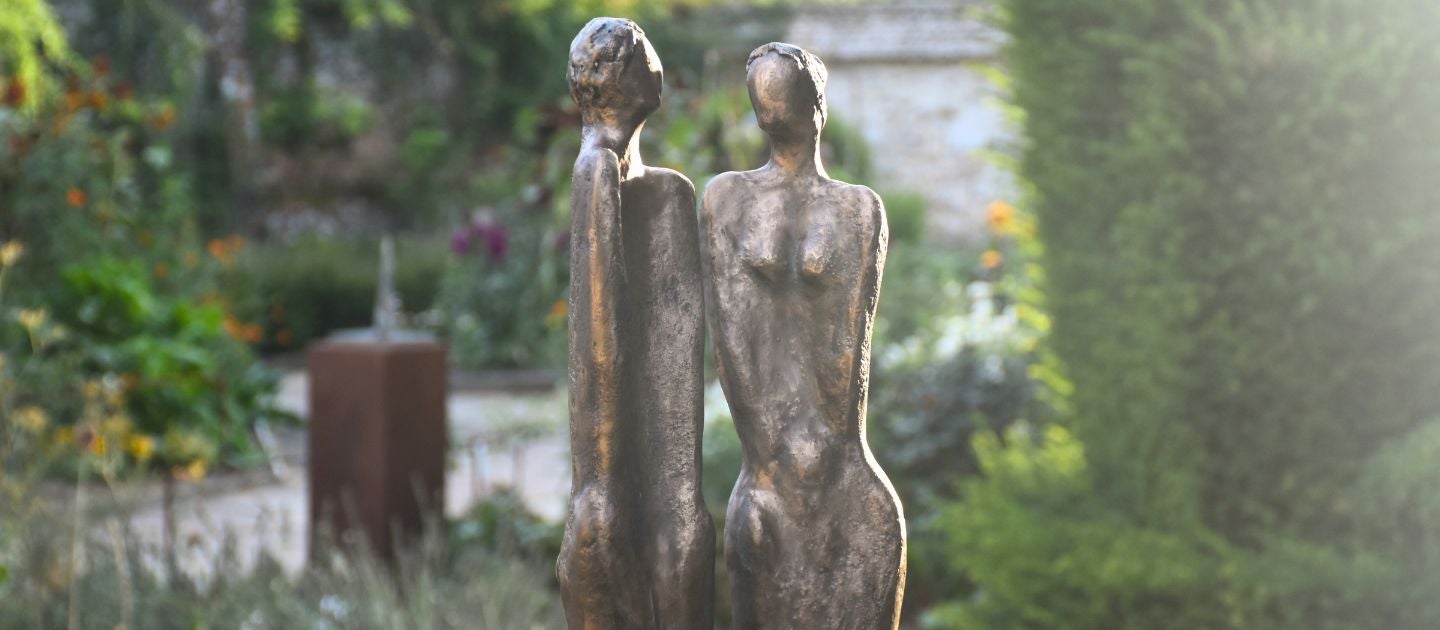 A close up of two figurines, a man and a woman against a blurred background of clipped green topiary and bright coloured flowers