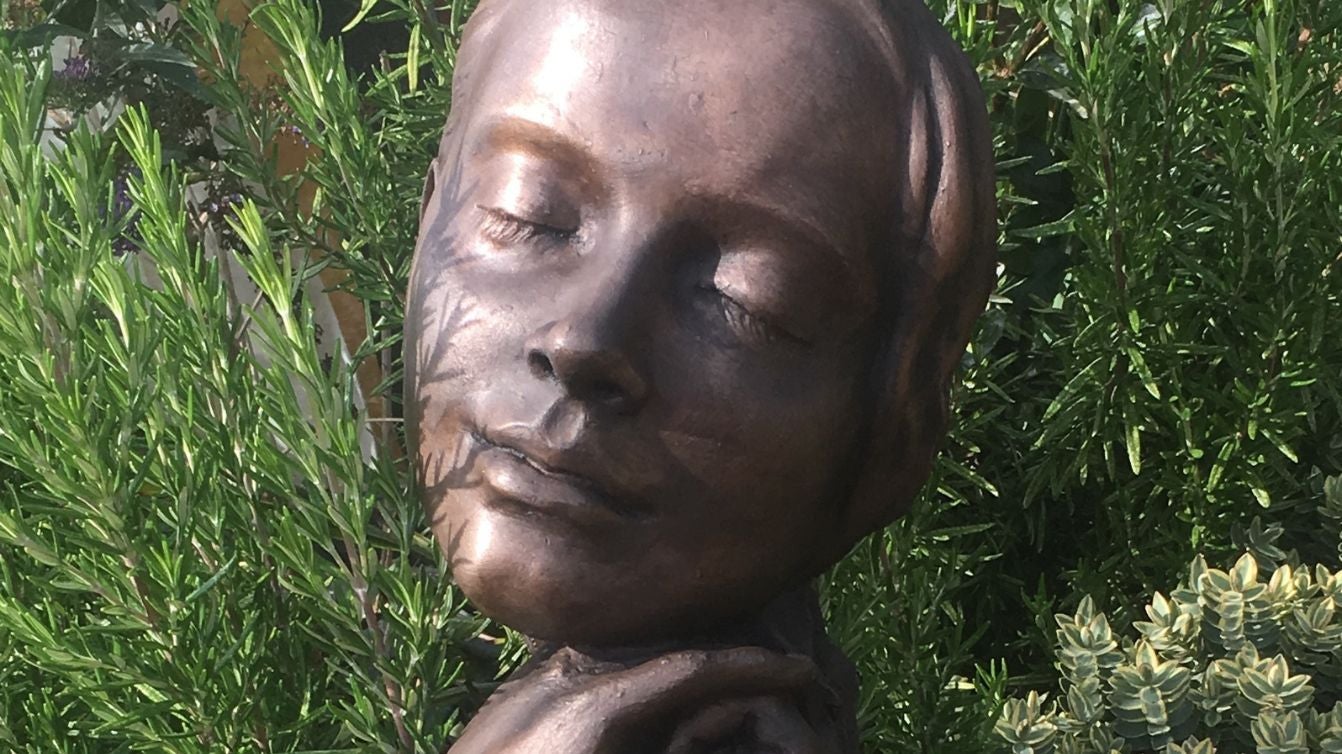 A close up of a bronze statue of a woman's face, set against green plants and hedges