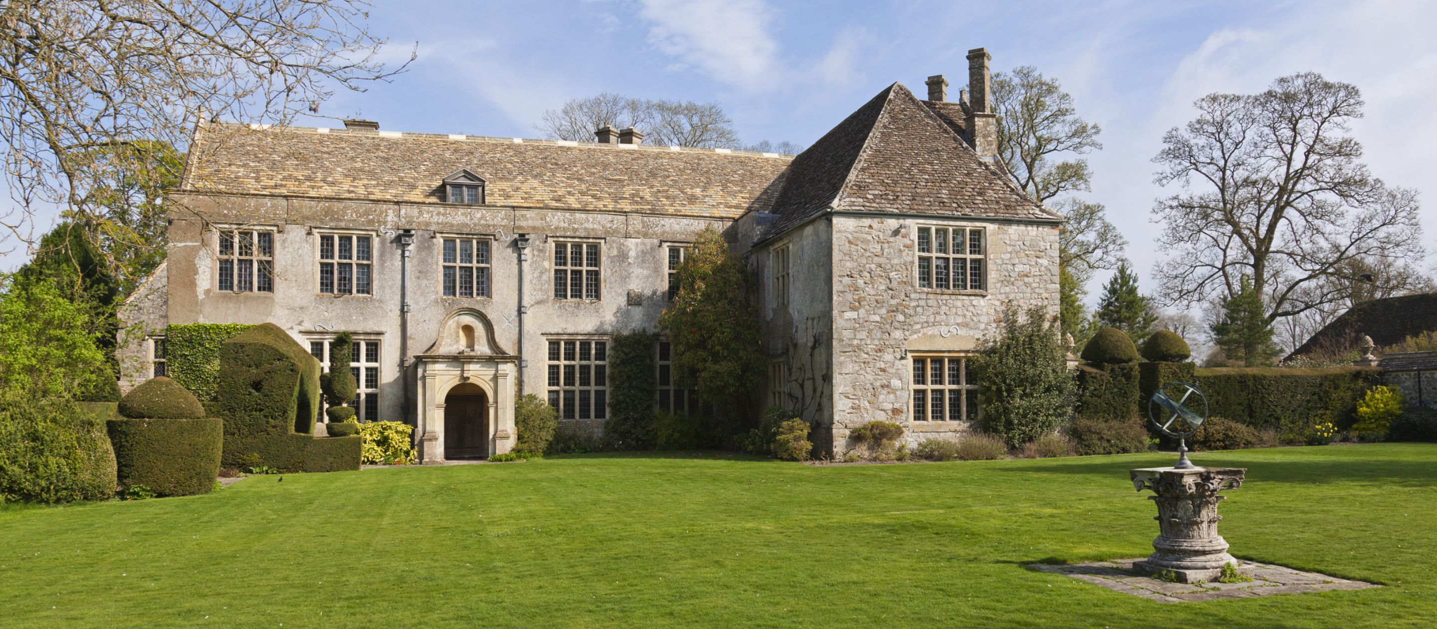 A wide landscape shot of the south front of Avebury Manor, with a lush green lawn in front of the house and a blue sky in the background.