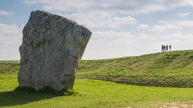 A large standing stone at Avebury in Wiltshire, with a group of people walking on the grass-covered henge bank in the background