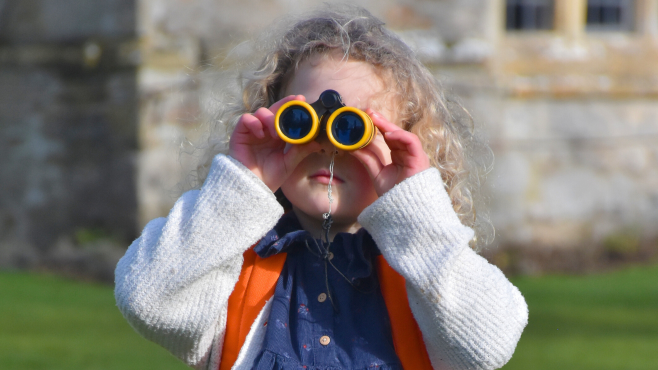 A girl looking through a pair of binoculars with green grass and part of Avebury Manor visible in the background.