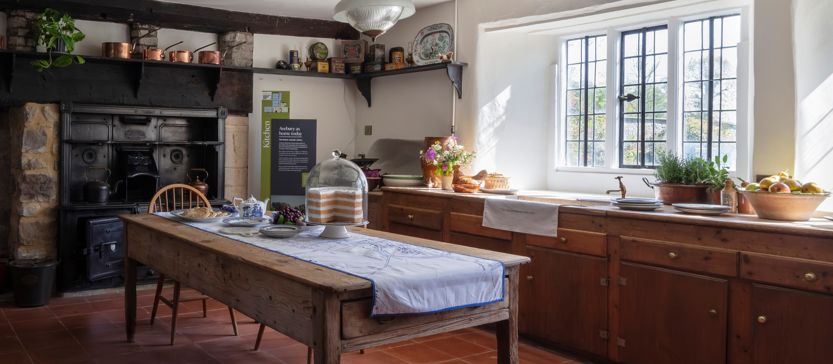 A wide shot of the kitchen at Avebury Manor, with a table laid with cups, teapot and large cake, with flowers and brass pots and pans arranged around a large stove
