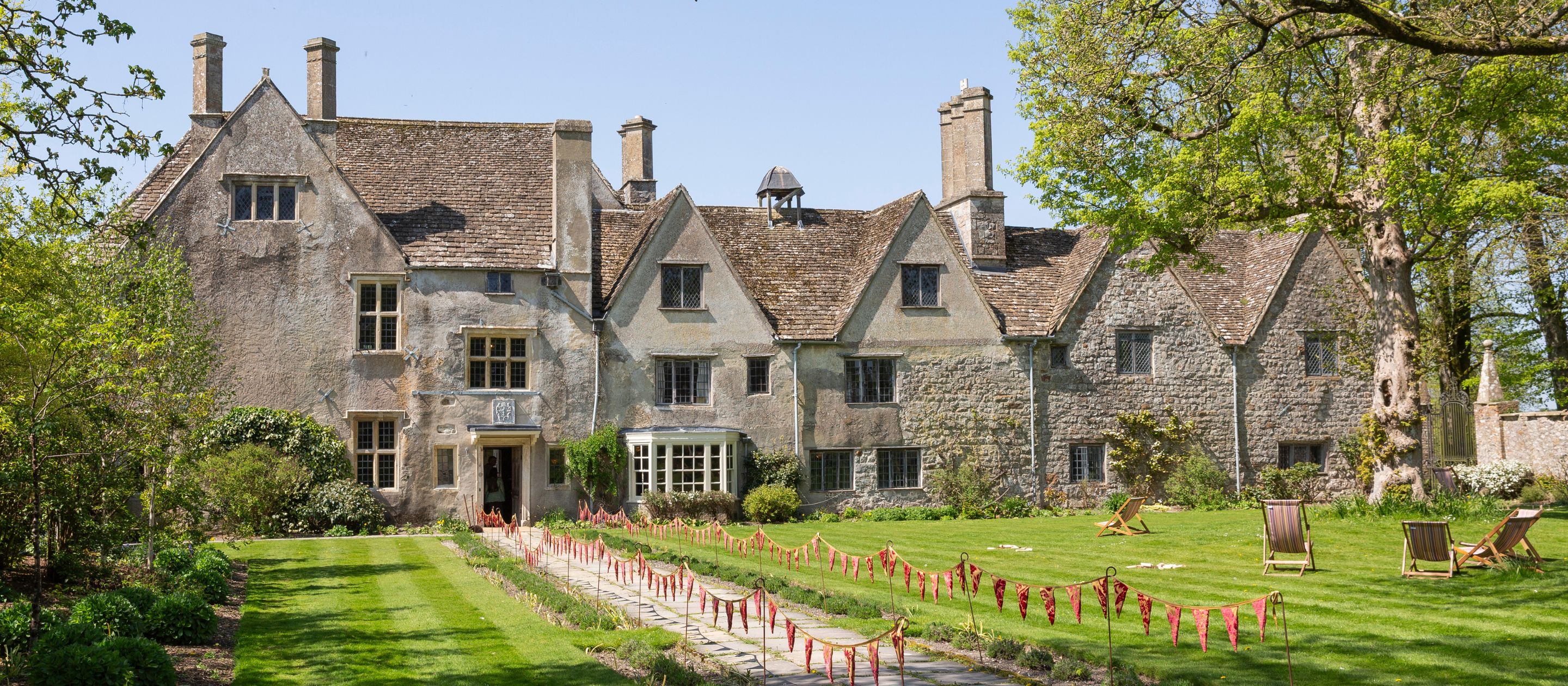A wide photo of Avebury Manor taken from the end of the Lavender Walk on the East Lawn, flanked by green trees and a bright blue sky.