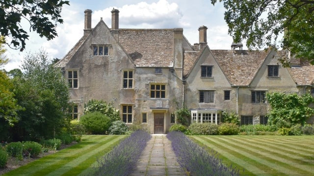 A landscape shot of the East Garden in Avebury Manor Garden showing flower beds on the left and a cut lawn on the right. Purple lavender lines both sides of the footpath through the middle of the photo, with the path leading to the door of the 16th Century Avebury Manor house.