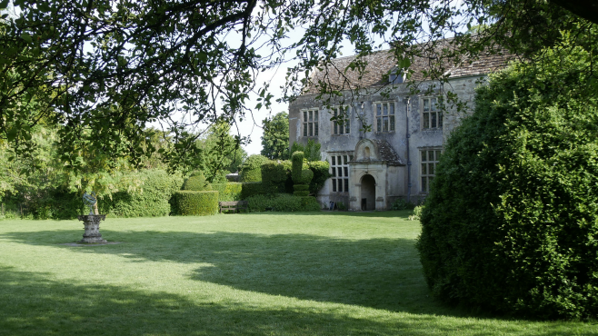 A landscape photo of the South Lawn in Avebury Manor Garden, flanked by large green hedges and trees and with the 16th-century manor visible in the background.