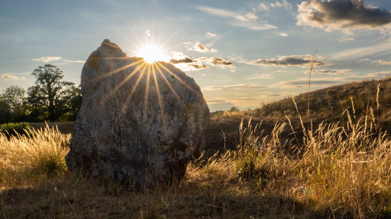 A standing stone in the NE sector at Avebury henge, with the setting sun visible behind the stone and henge bank.