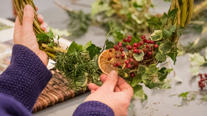 Close-up of hands in a blue sweater creating a decorative wreath. The wreath features fresh green foliage, red berries, rosemary sprigs, and a dried orange slice being attached to the arrangement."