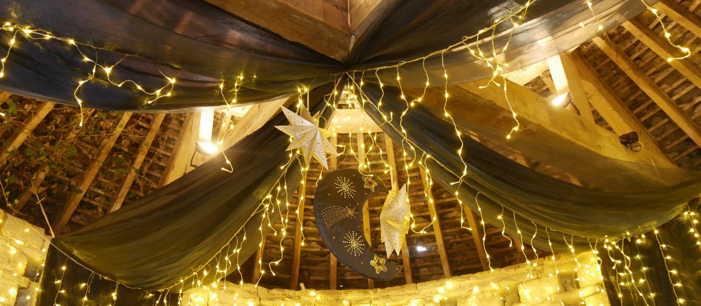 The timber frame ceiling of Avebury's 16th Century Dovecote decorated with black drapes, hanging paper star ornaments, warm fairy lights, and a crescent moon decoration featuring golden star and comet designs.