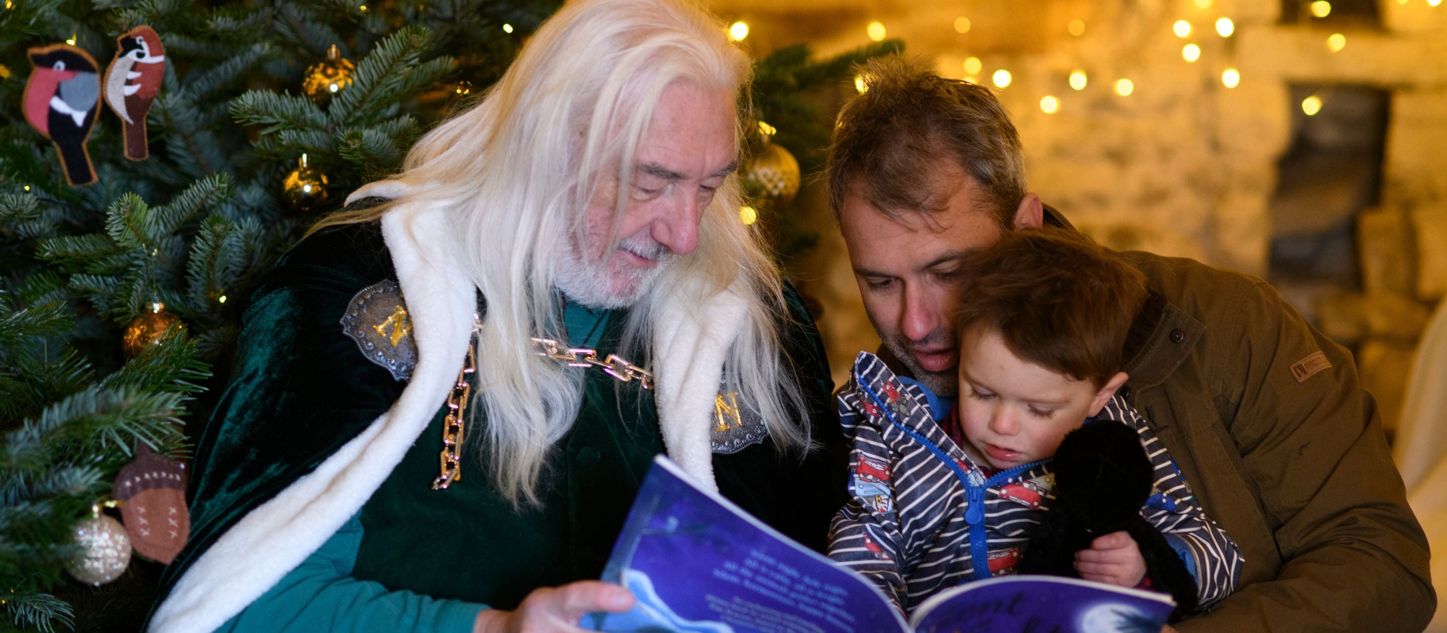 St Nicholas with long flowing white hair and a green cape reading a story to a boy sat on his father's lap in the Dovecote, surrounded by trees and glistening lights.