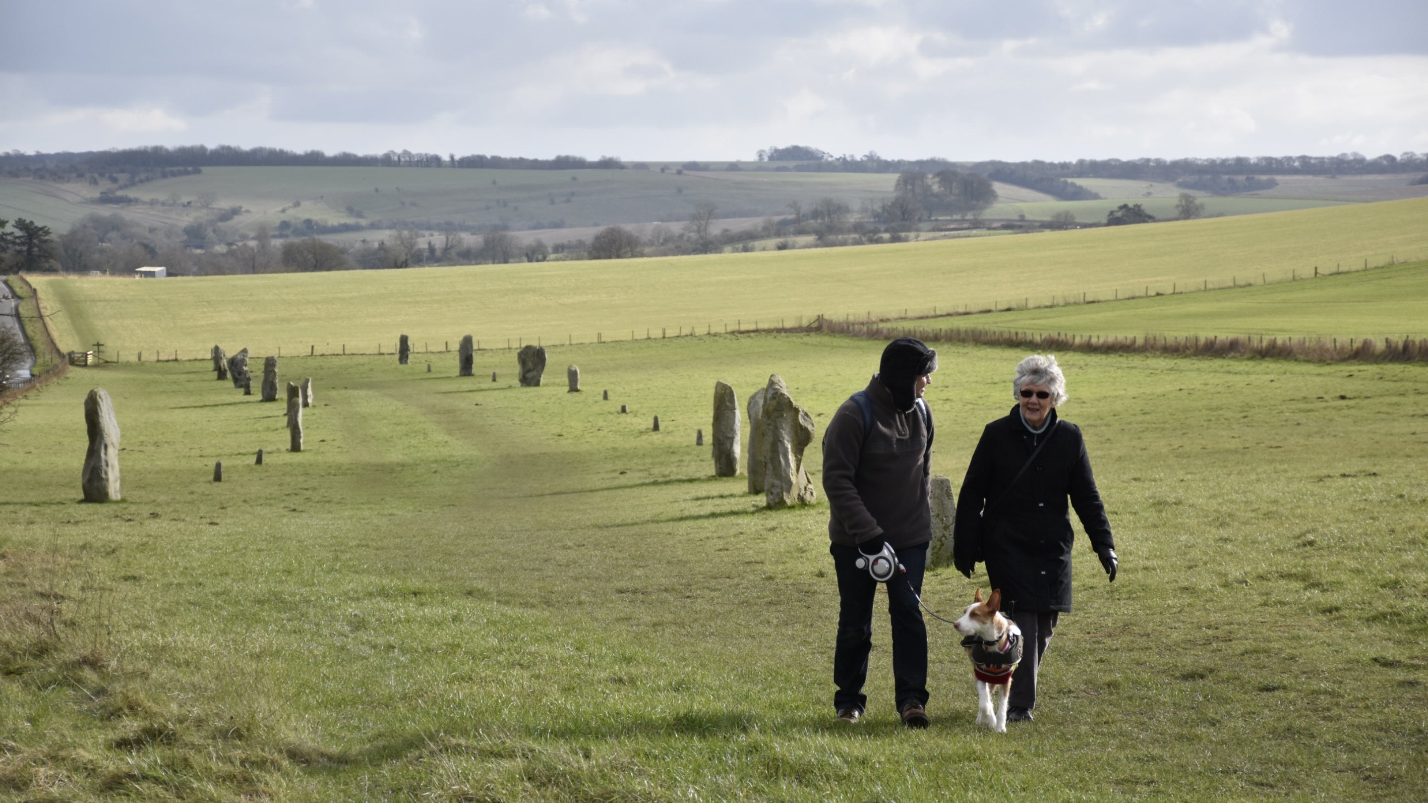 Two visitors with a dog on a lead walk up a grassy avenue lined by small standing stones either side of them