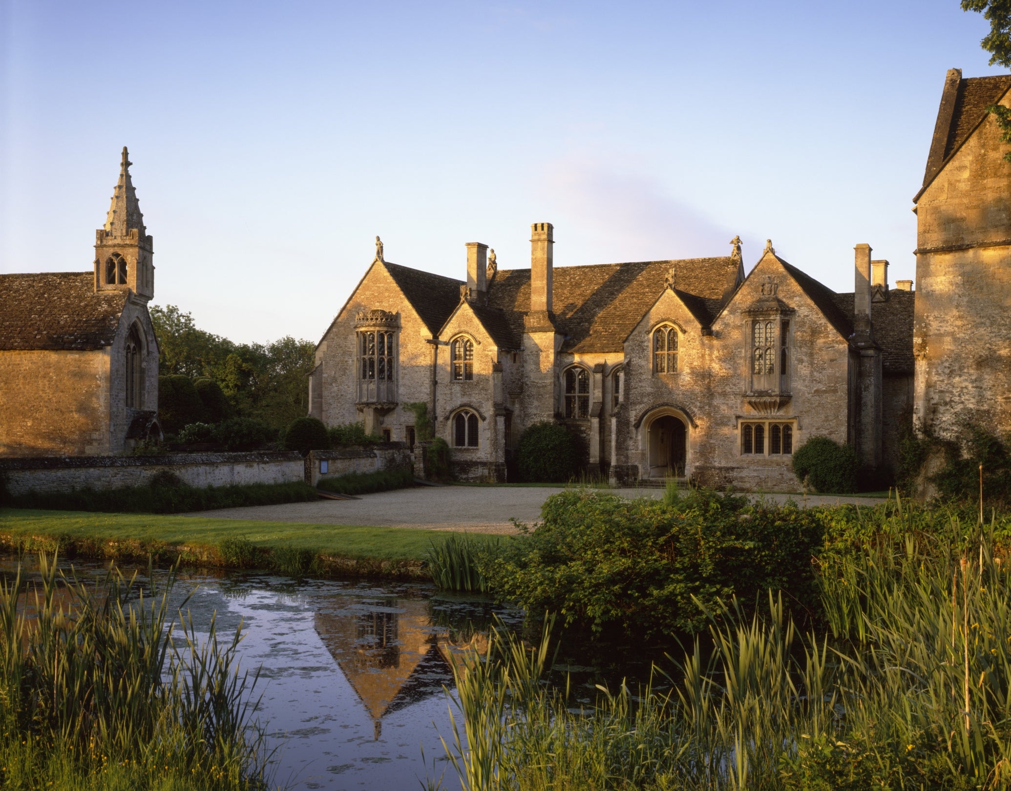 A view of the exterior of Great Chalfield Manor across the moat and forecourt, the sun is low in the sky and glinting off the roof.the
