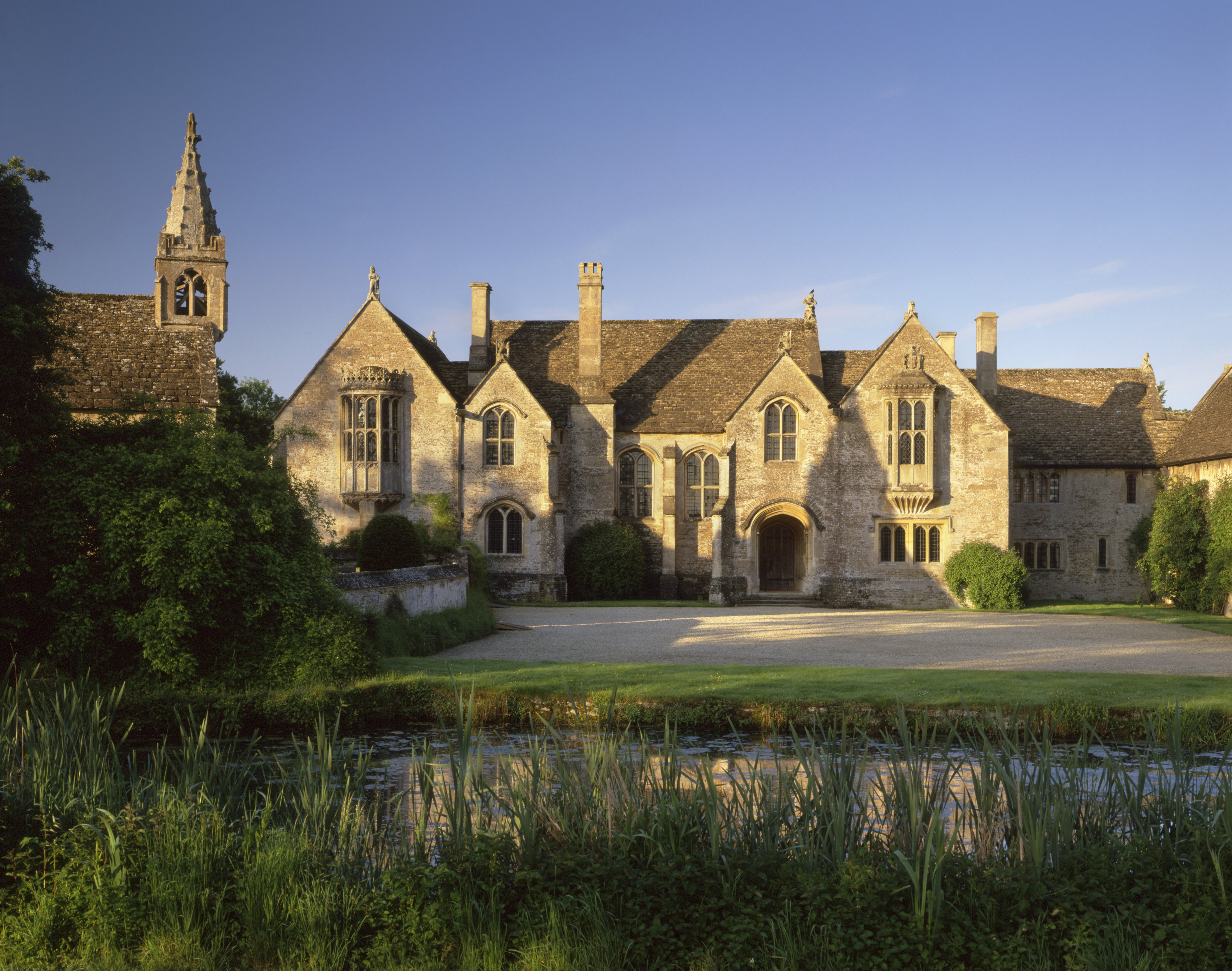 A view across the moat of the front and forecourt of Great Chalfield Manor, a medieval manor house in Wiltshire, with blue sky and low sun shining on the warm stone.
