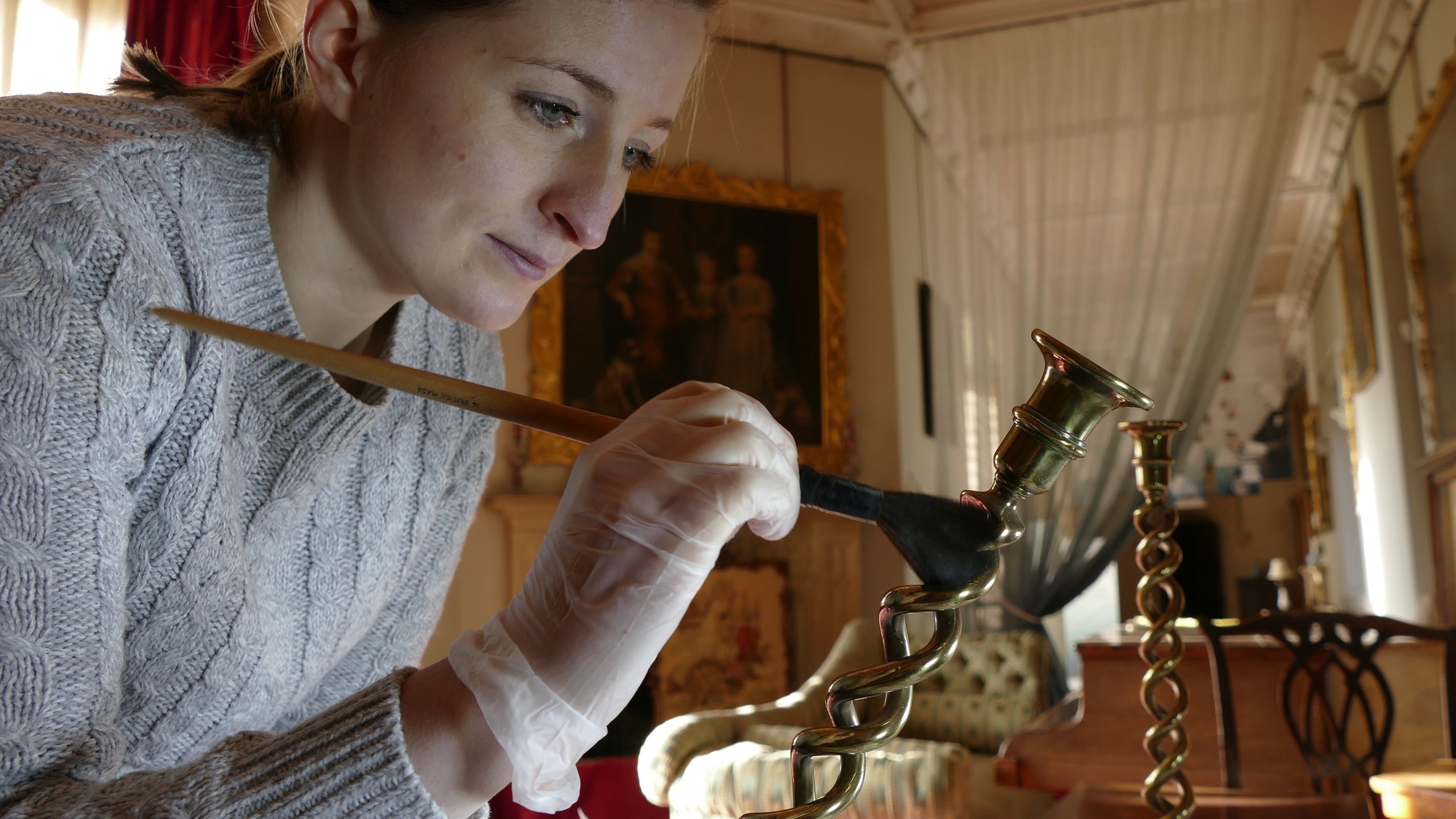 A conservator is cleaning a candlestick with a delicate brush in the South Gallery at Lacock, Wiltshire.