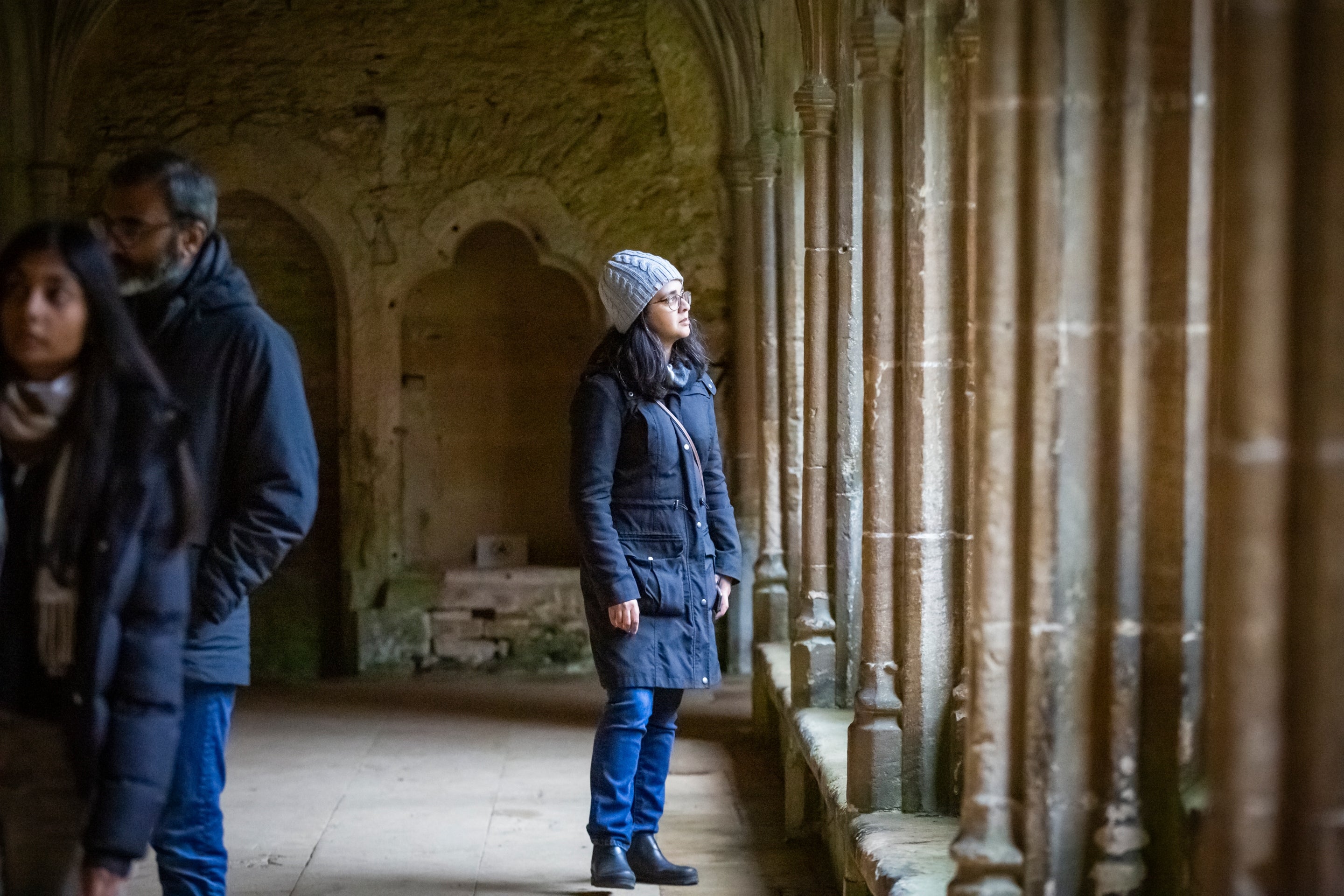 A visitor looks out of the window in the Cloister at Lacock during winter. They are wearing a warm hat and coat, and looking pensive.