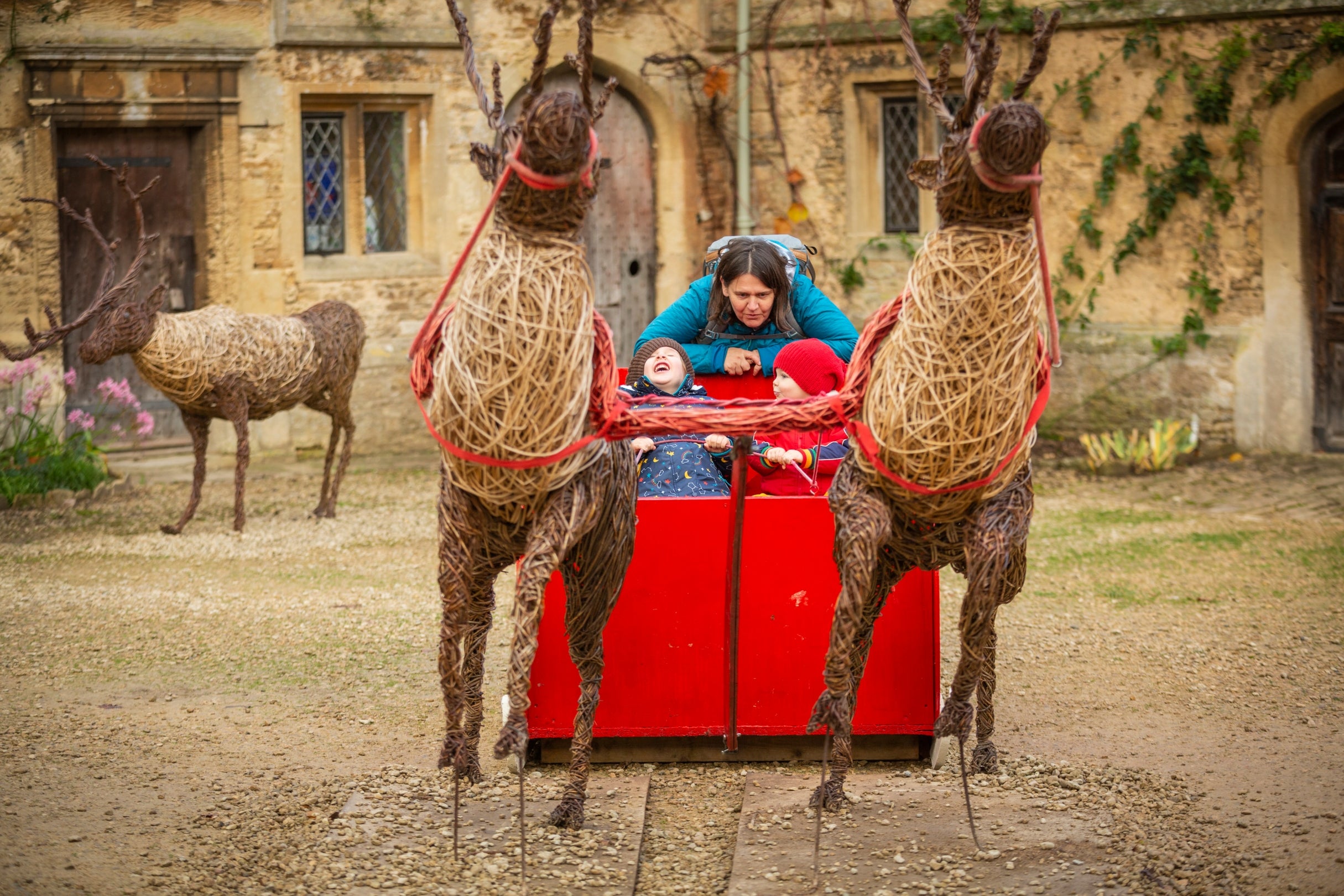 Two children sit in the reindeer sleigh at Lacock, they are laughing and their adult carer stands behind them joining in the fun.