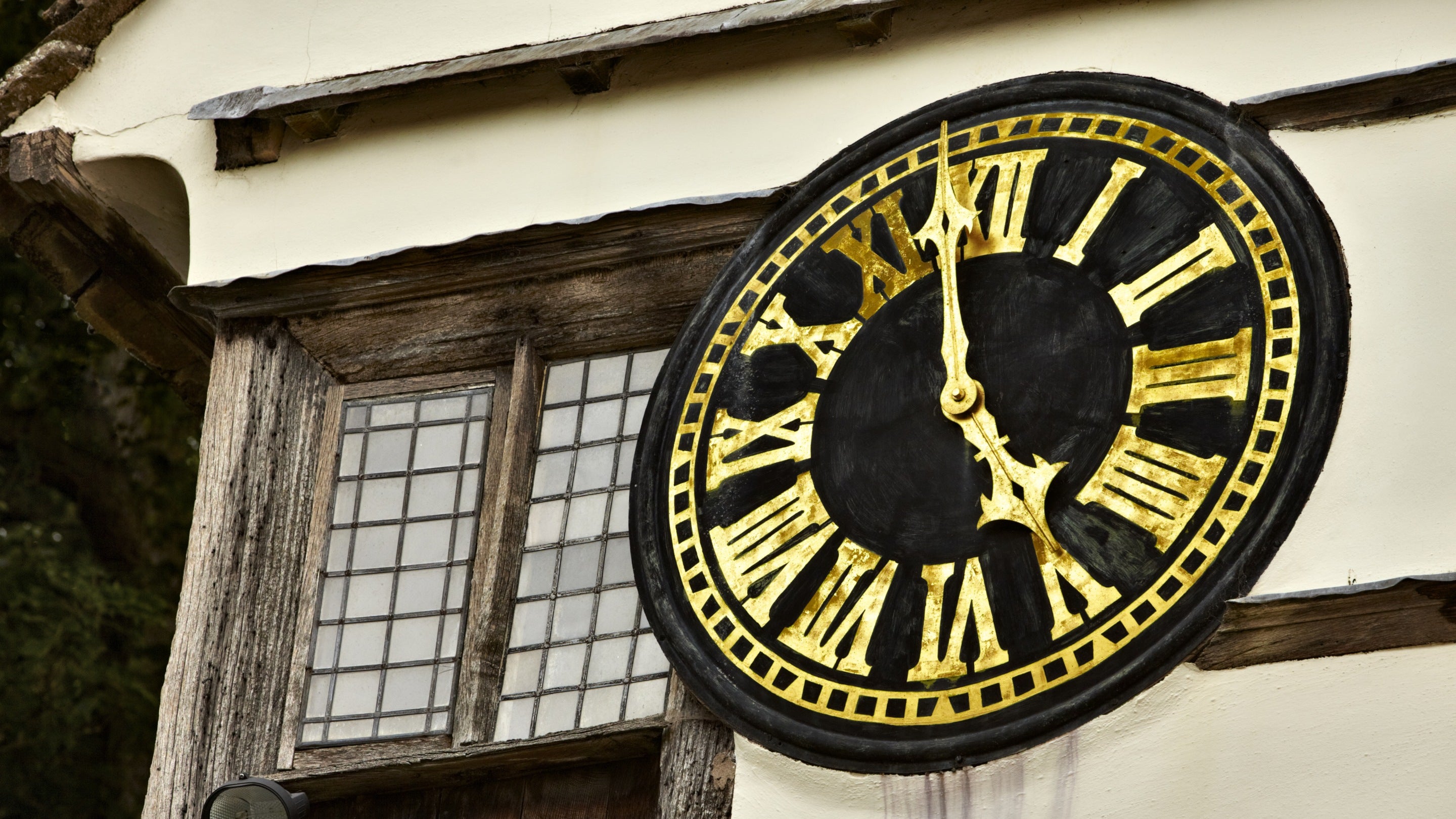 A large black clock face with gold roman numerals and hands is shown on the side of a historic building at Lacock