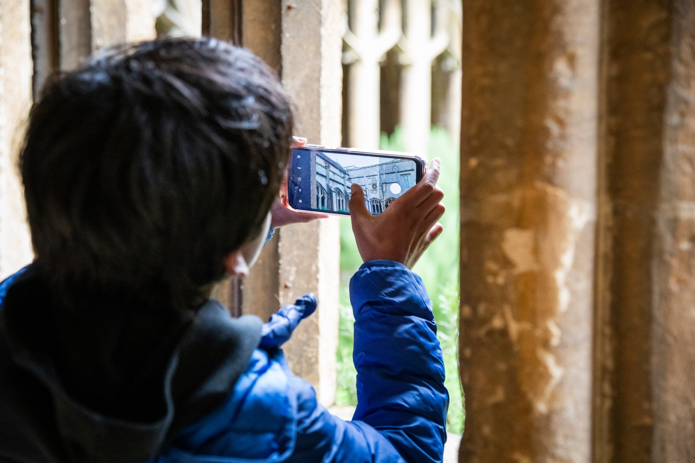 A child gets ready to snap a photo on their phone through the window of the Cloister at Lacock, they use their thumb to focus the image which captures the stone walkway across the Cloister Garth.