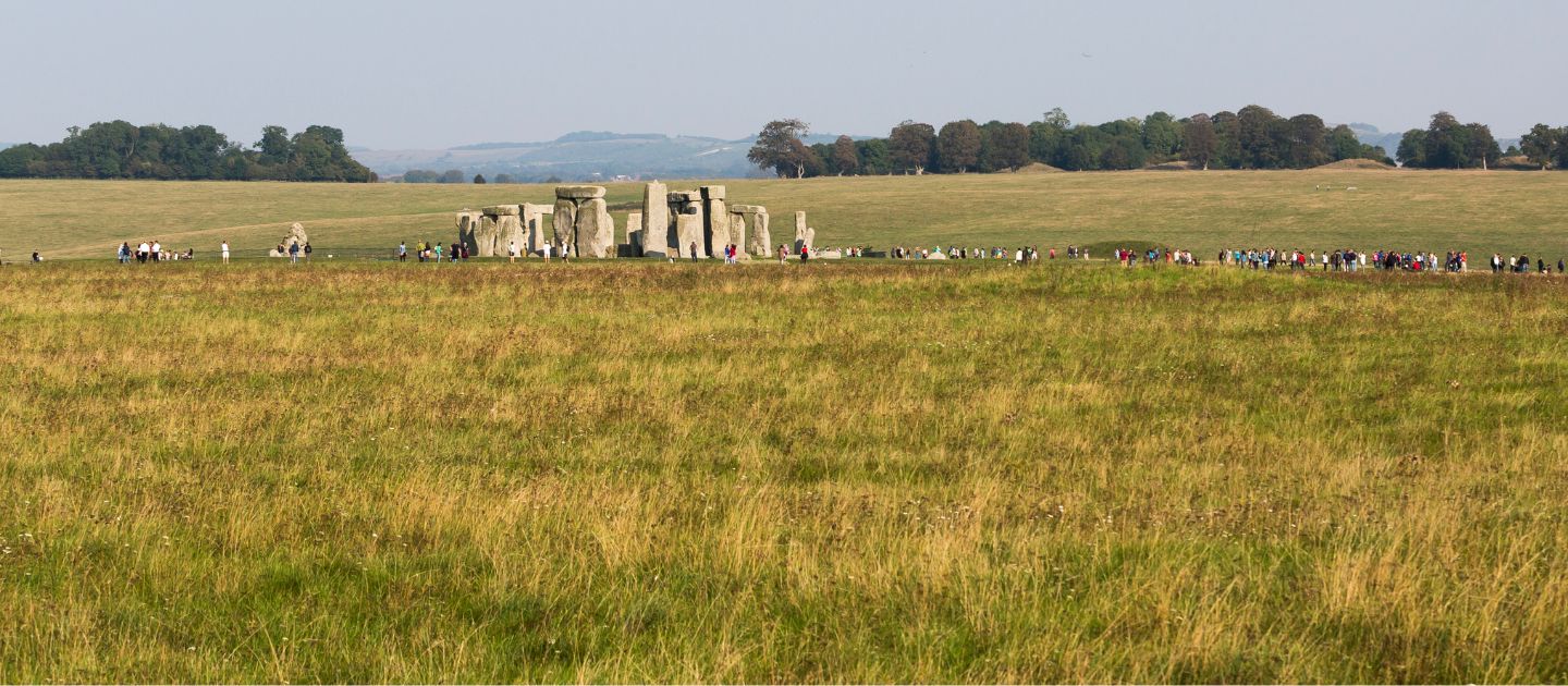 A wide landscape shot across a chalk grassland field with the Stonehenge monument and visitors visible in the background
