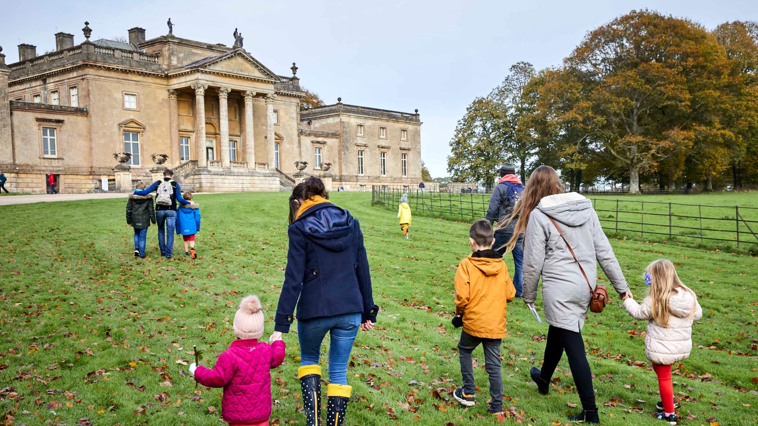 Children and adults wearing hats and coats walk up a hill in front of the house at Stourhead