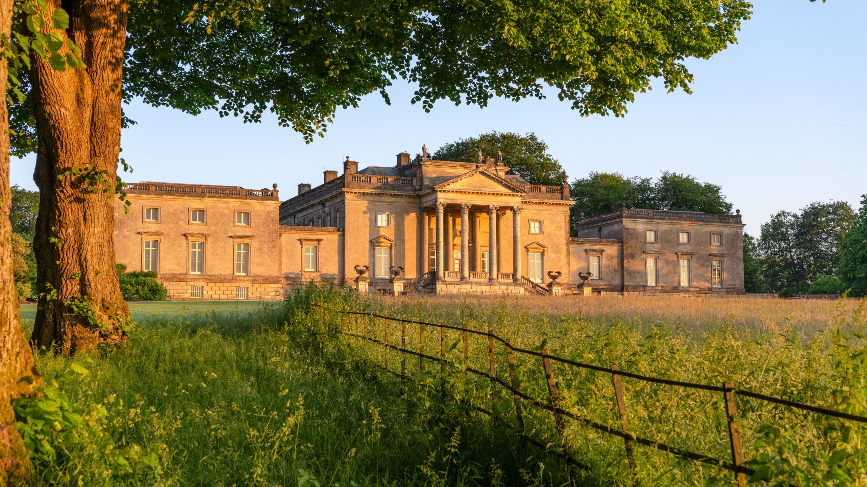 View across a meadow to the golden stone frontage of the house at dawn, Stourhead, Wiltshire