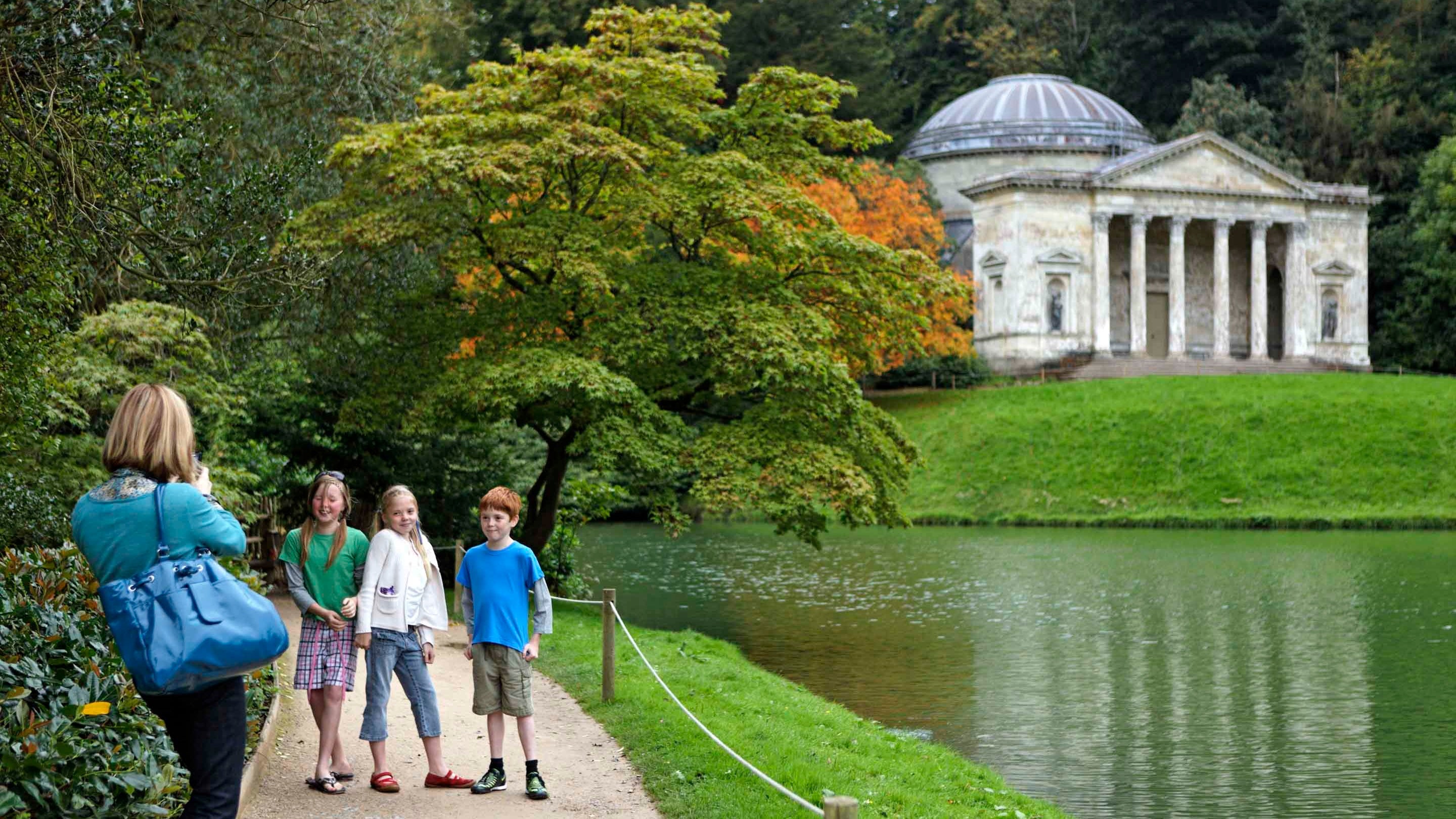 Three children having their photo taken by a woman in front of the lakes at Stourhead with the domed Temple of Apollo in the background