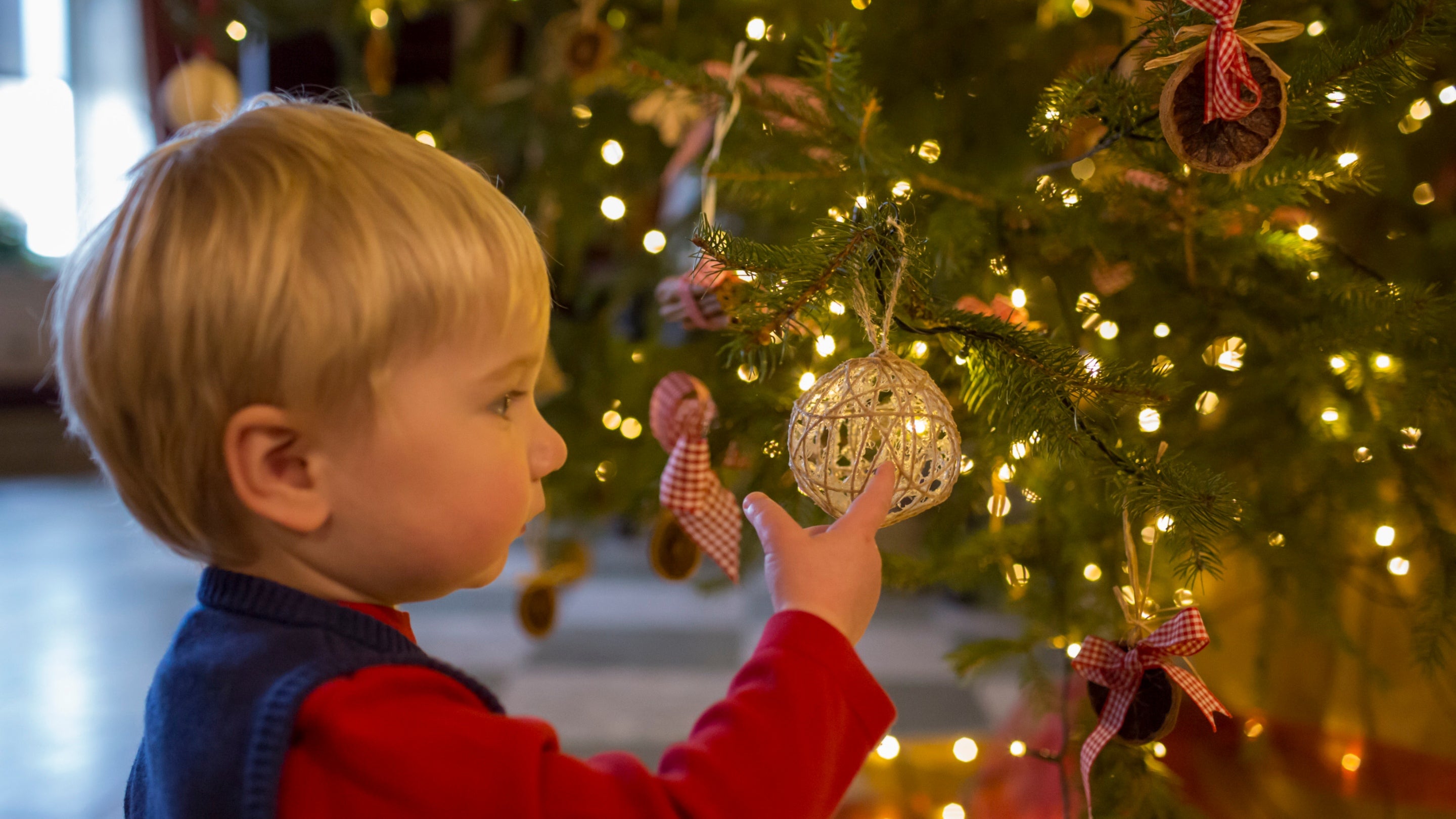 A young boy wearing a blue knitted tank top points at a natural woven bauble on a Christmas tree glowing with fairy lights