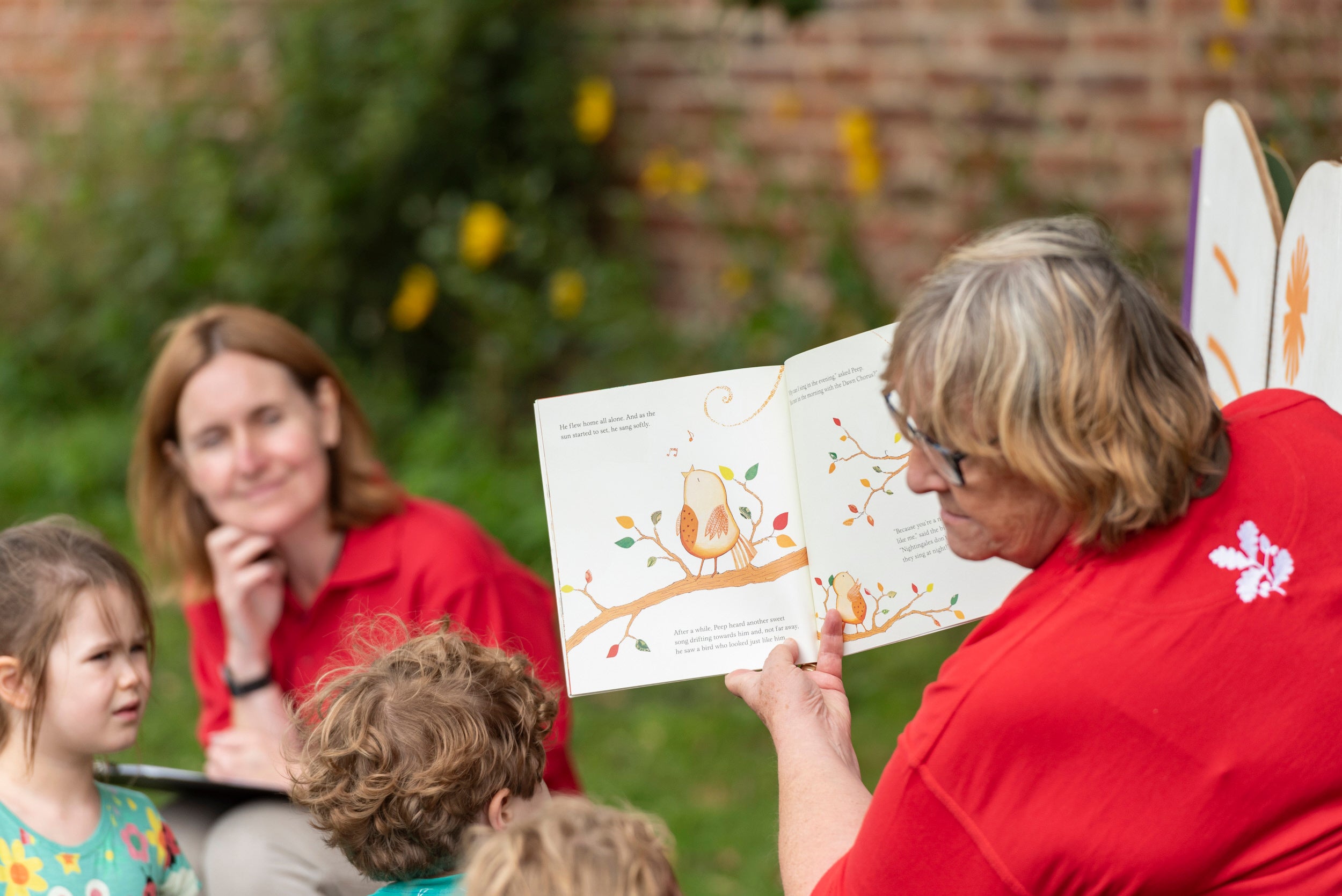 Volunteer reading a story from a book at a summer event with children sat on the grass listening