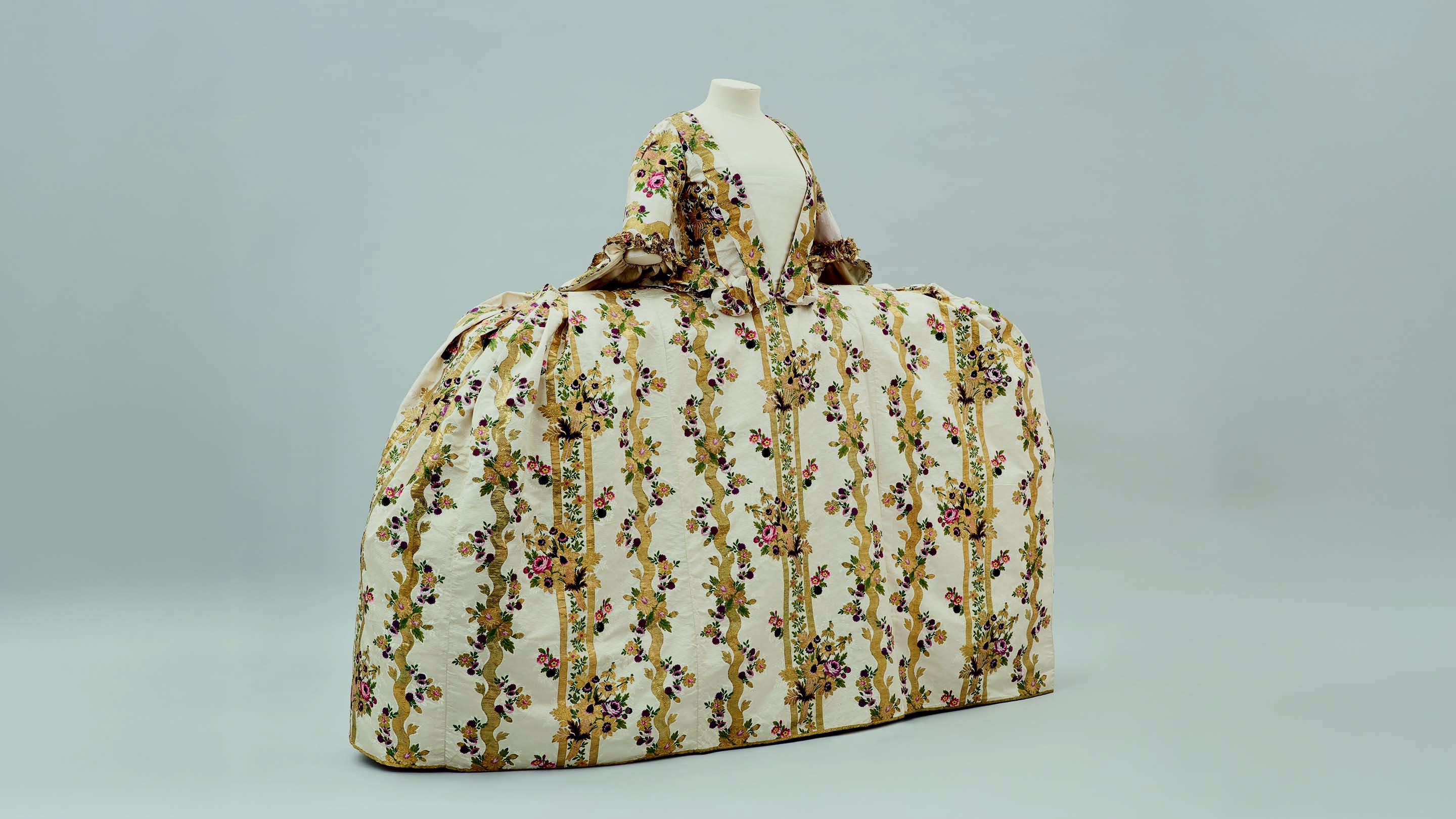 A very wide 18th century dress made from cream silk brocade with a luxurious design of tumbling floral bouquets against stripes and meanders of gold thread.
