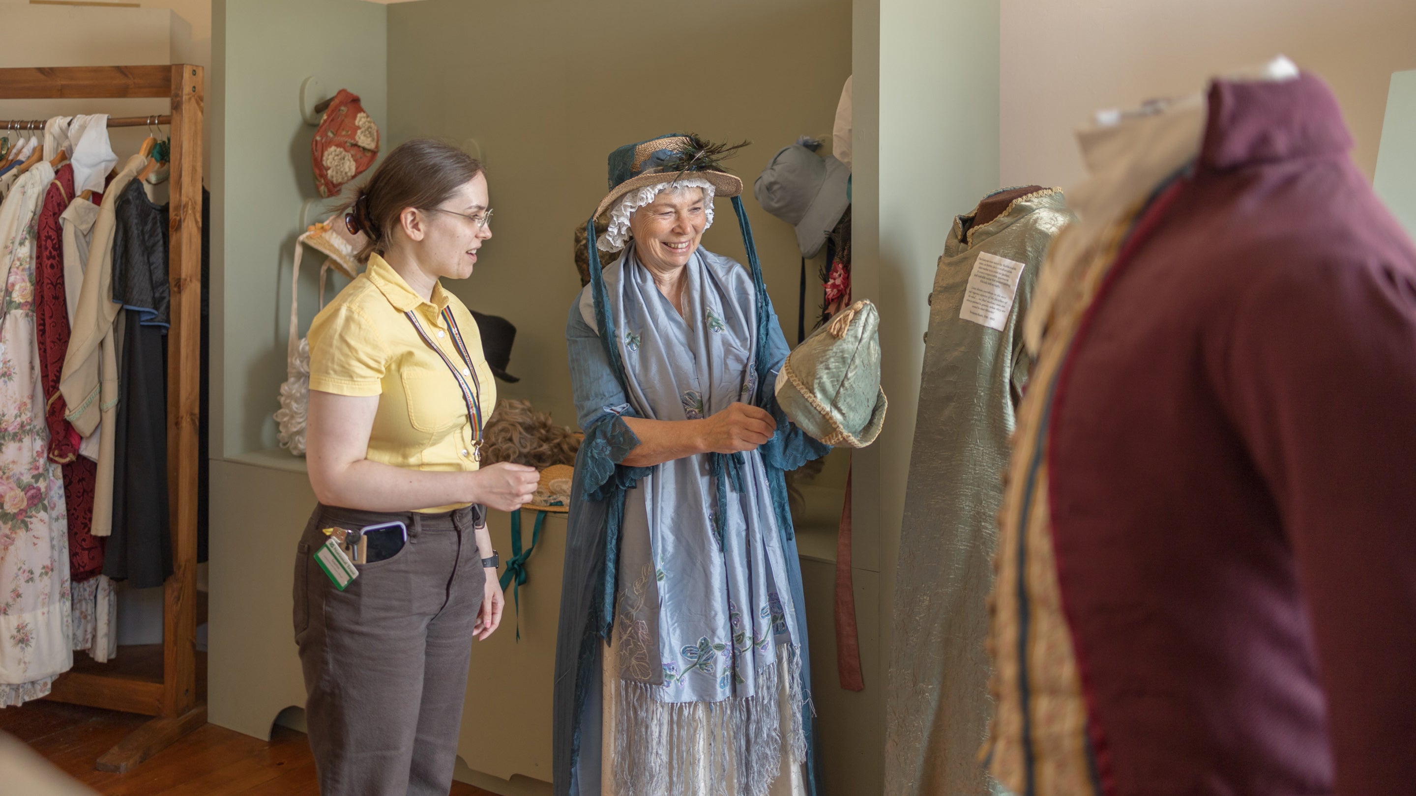 A volunteer dressed in Georgian costume wearing a bonnet draped in ribbon and blue silk scarf. The volunteer is showing a member of the House team a hat and they are smiling.