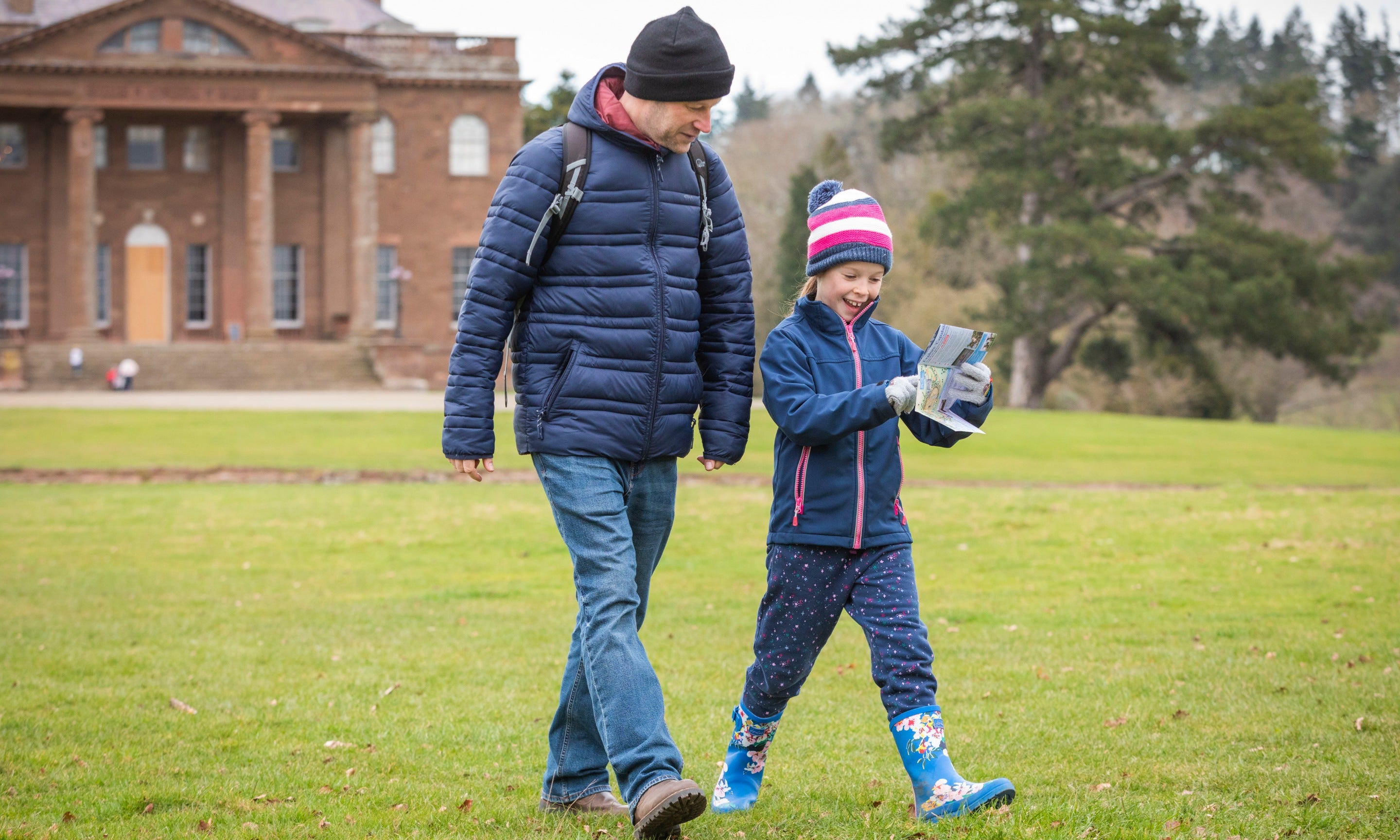 A man and a girl walking while looking at a map, dressed in winter clothes and wellies, with Berrington Hall in the background