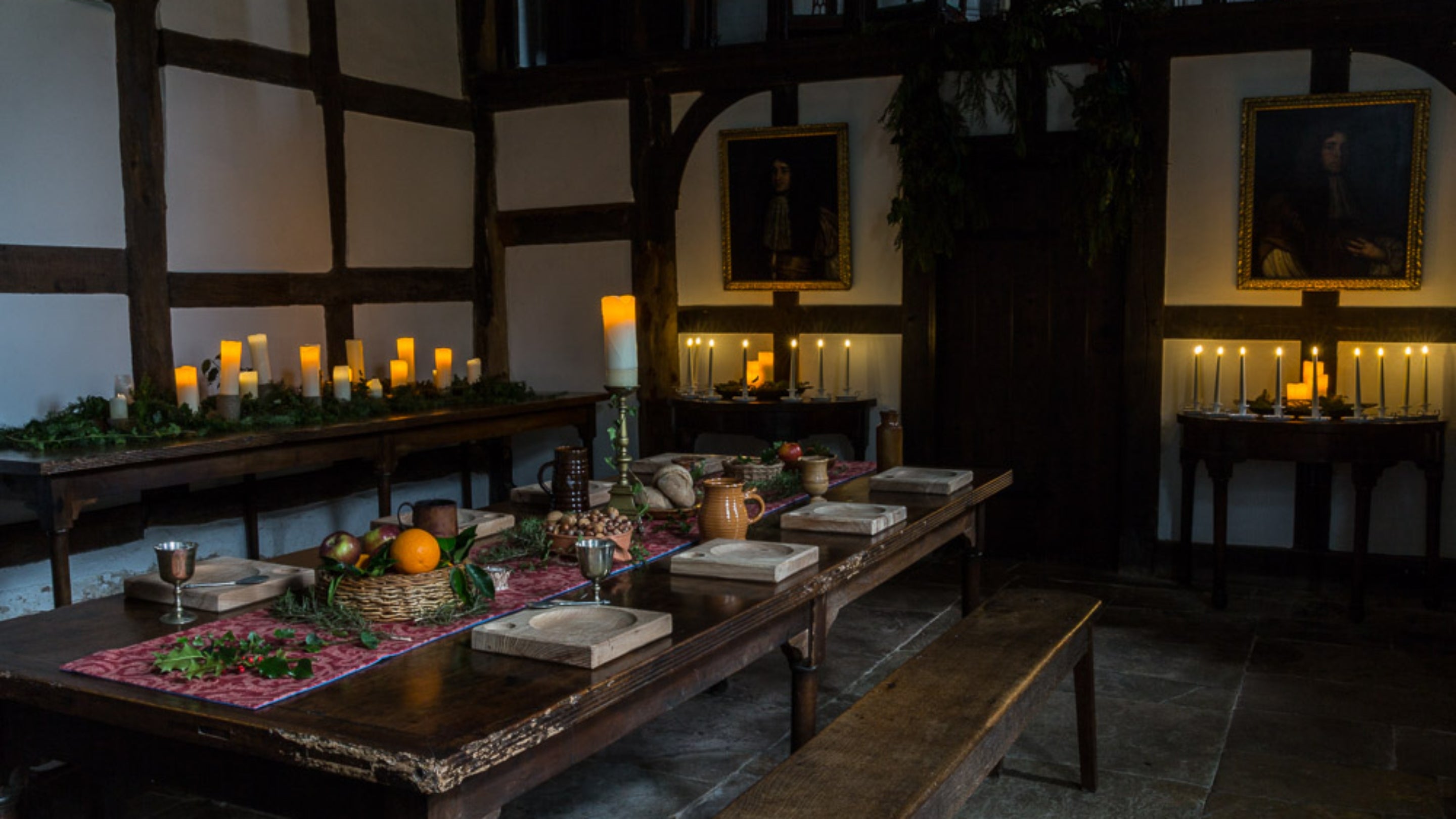 The Great Hall at Brockhampton, Herefordshire is decorated with seasonal greenery on a red central tablecloth, with lots of lit white pillar candles on side tables around the room