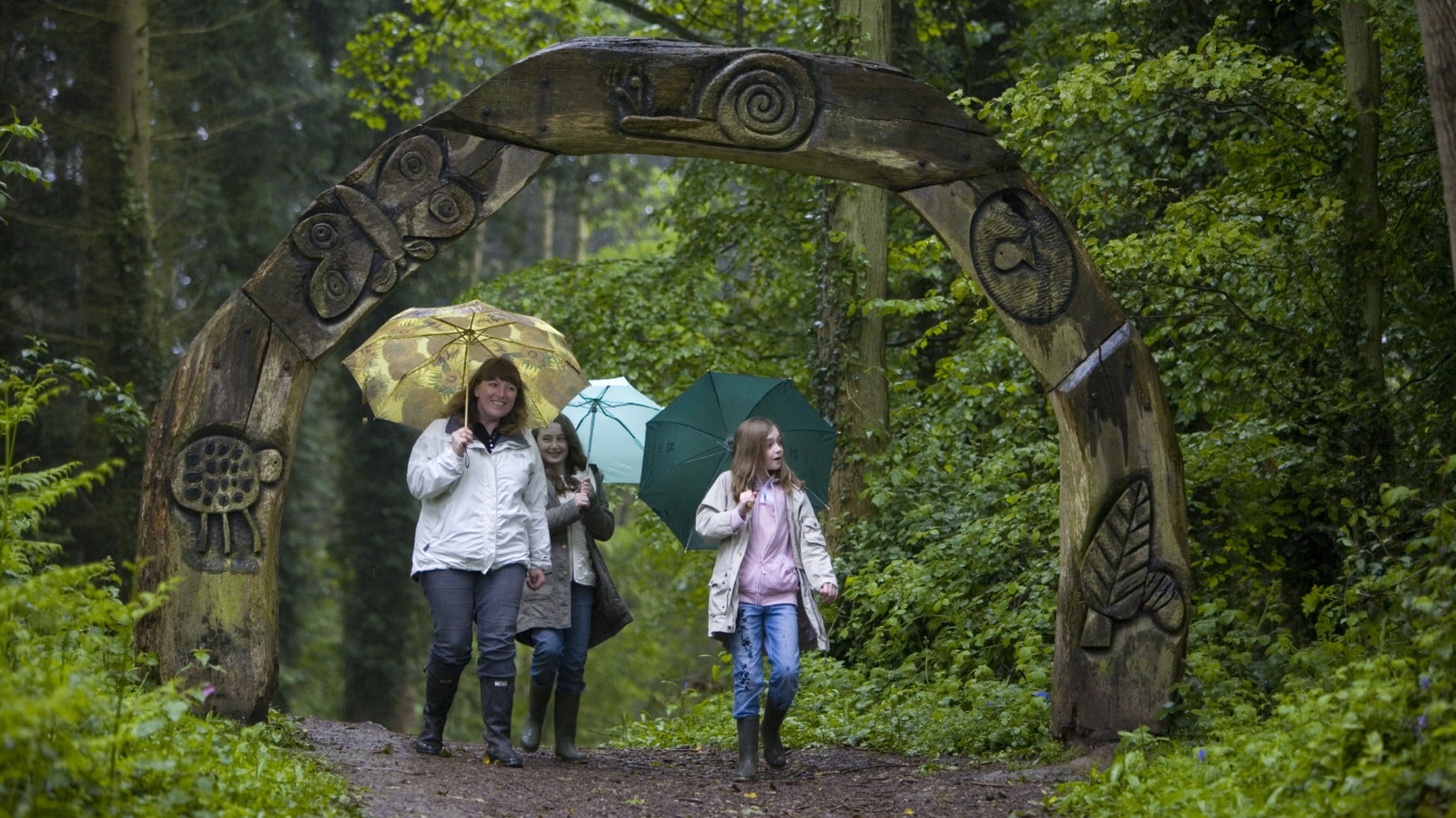 A family wearing raincoats and holding umbrellas walking beneath a wooden archway carved with leaves, snails, butterflies and other nature images at Brockhampton