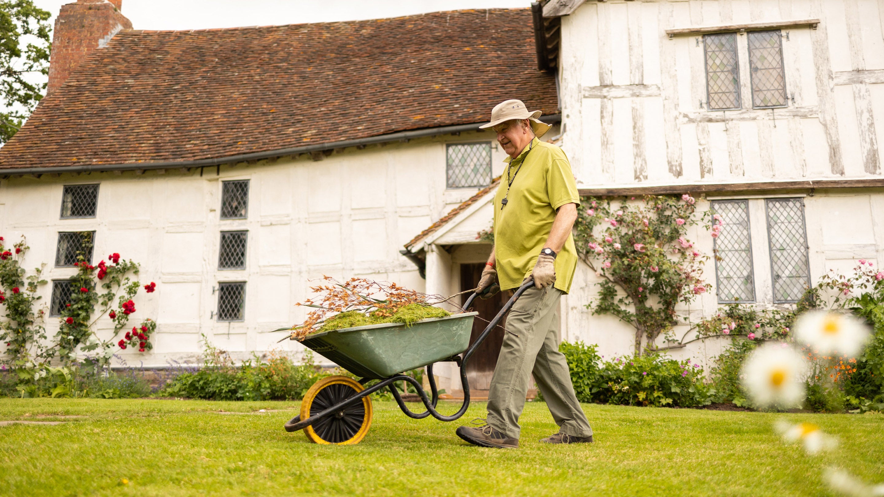 Volunteer gardener pushing a wheelbarrow wearing a green top and smiling, in front of a timber framed medieval manor house.