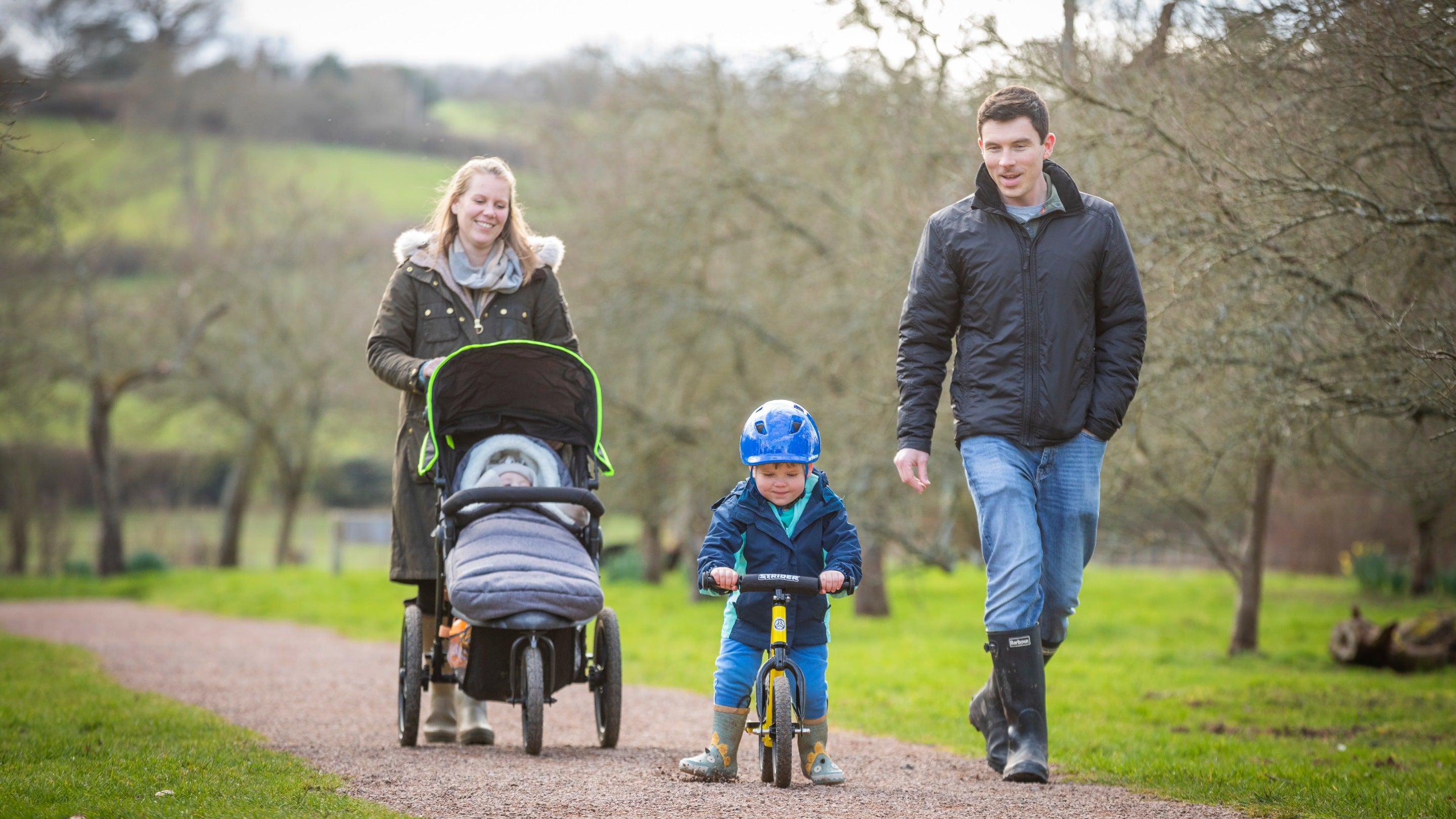 A family walking along a brown path that leads through the orchard. The woman is pushing a pushchair, and the man walks alongside a young boy on a small bike.