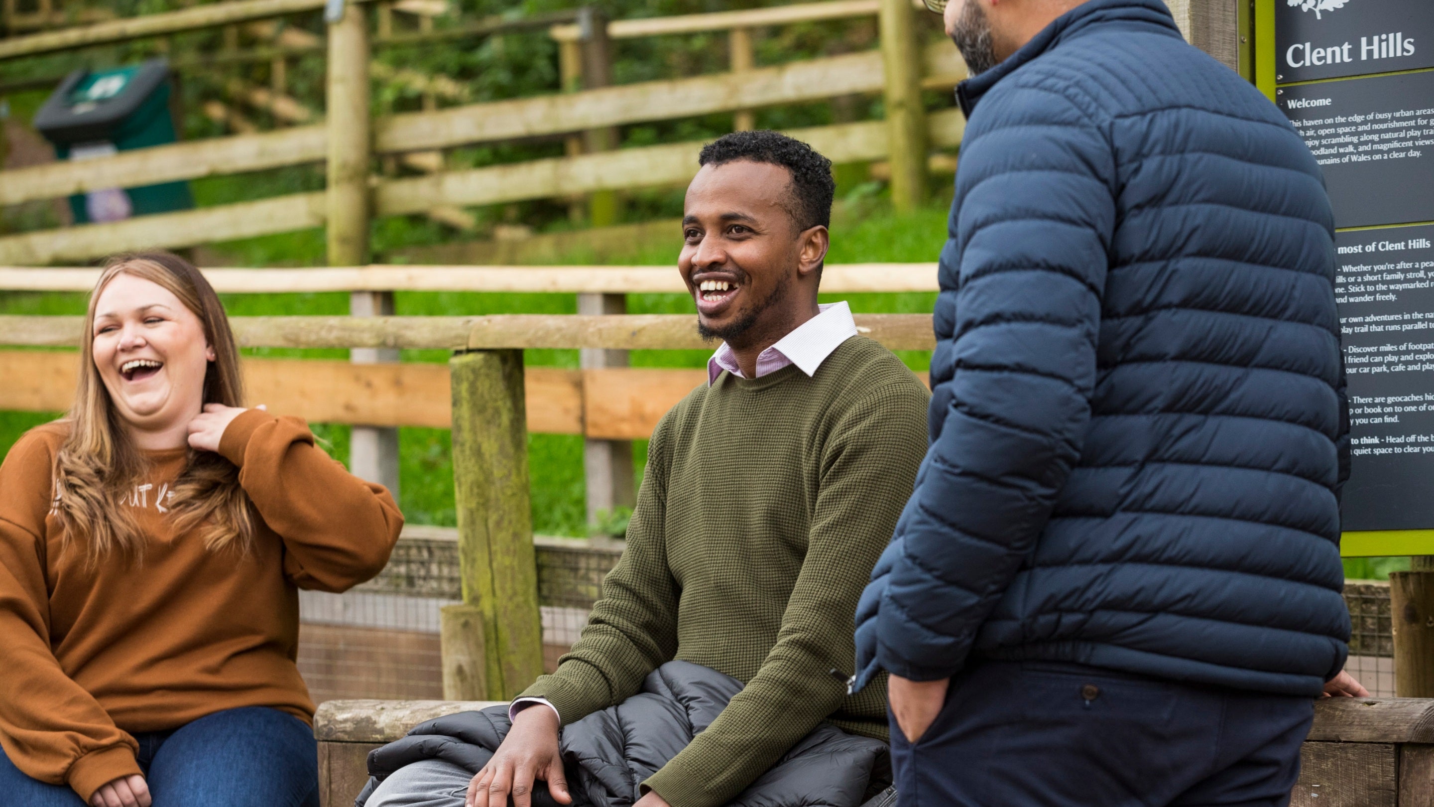 A close-up of two visitors sitting on a wall and laughing, with another visitor standing in front of them at Clent Hills