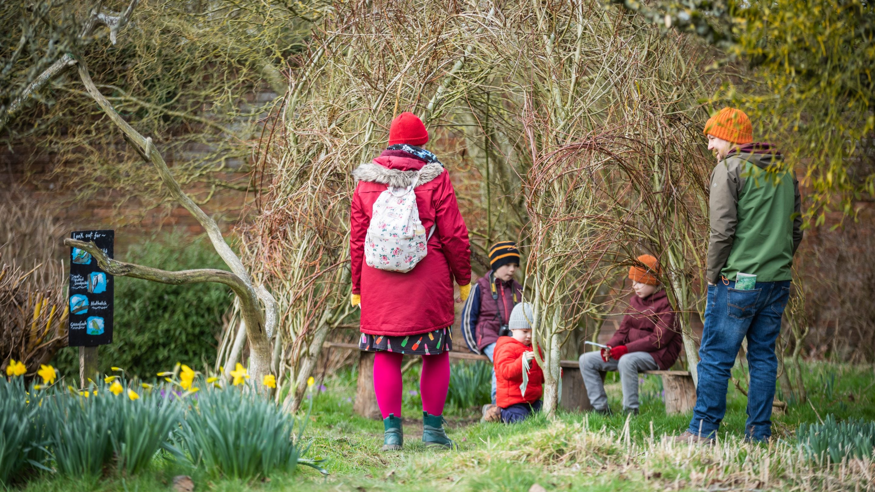 A family sitting on a bench under a tent-like structure weaved out of willow in the walled garden.
