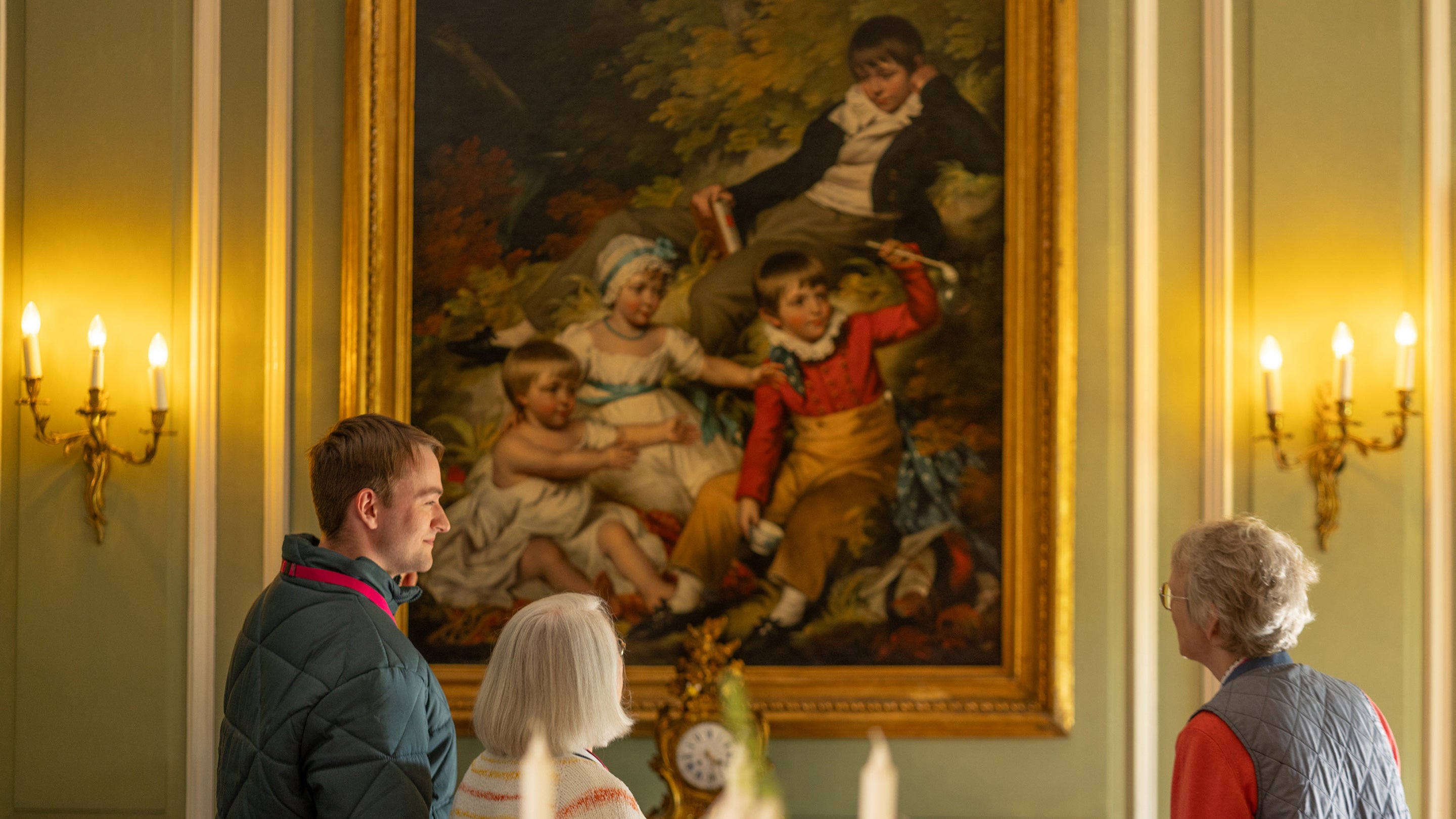 Volunteer smiling at visitors in front of a huge historical painting of children, hung on a sage green wall with candelabra's either side emitting warm light giving the whole picture a golden glow.